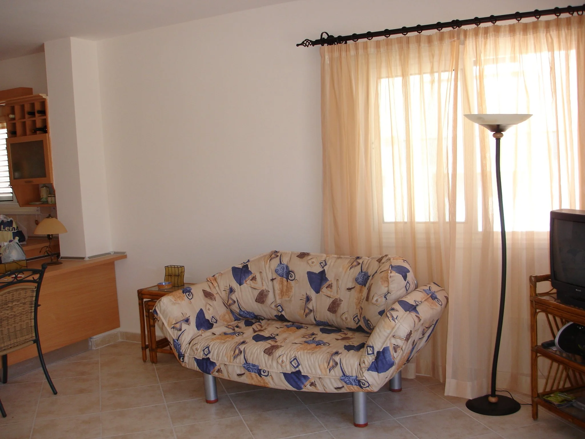 Living room with a floral-patterned loveseat, a floor lamp beside a window with beige curtains, a wicker TV stand with an older model TV, and a wicker chair with a small table.