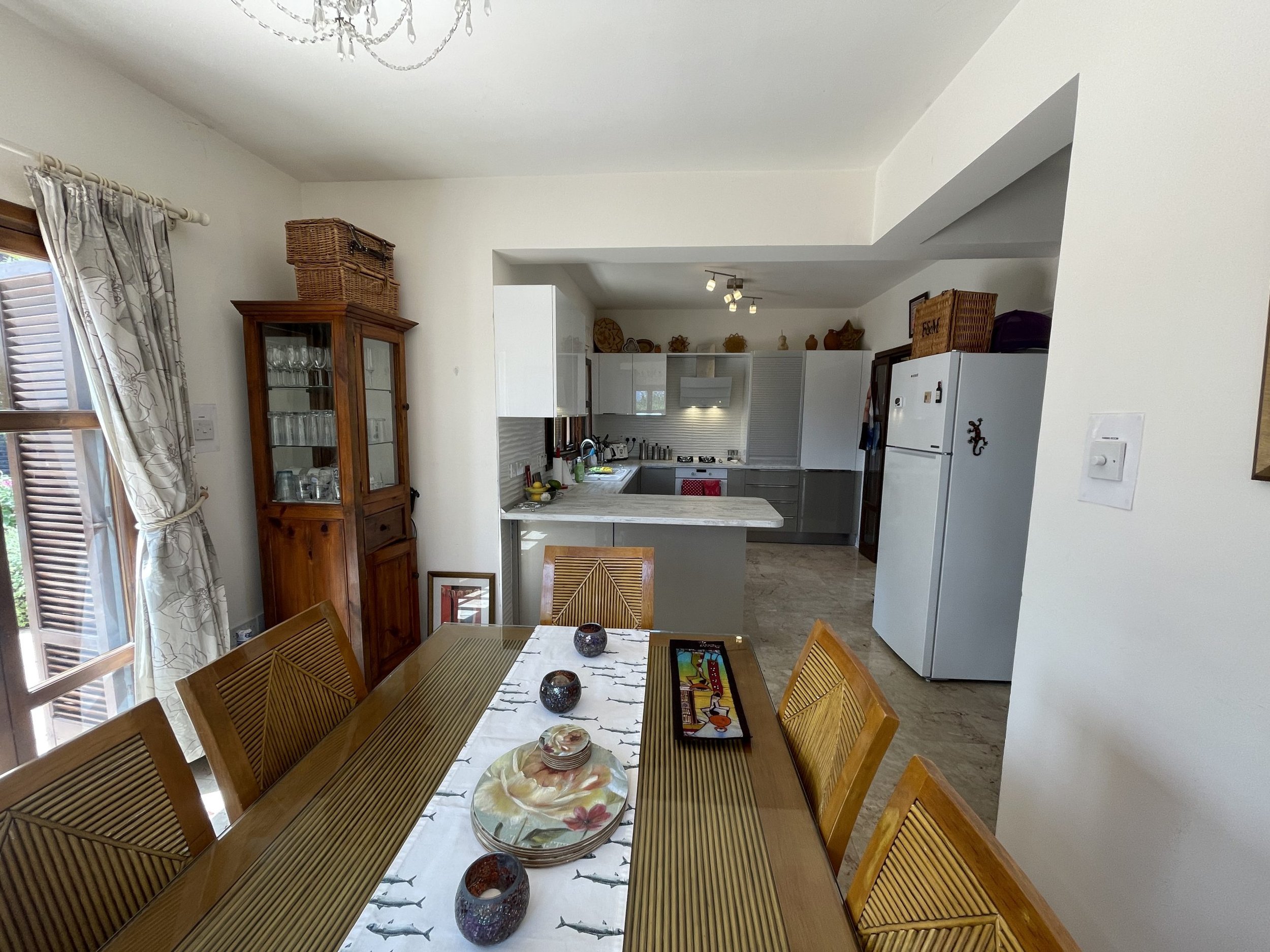 A dining room with a wooden table and chairs, a china cabinet, and a view into a kitchen with white cabinets and a refrigerator.