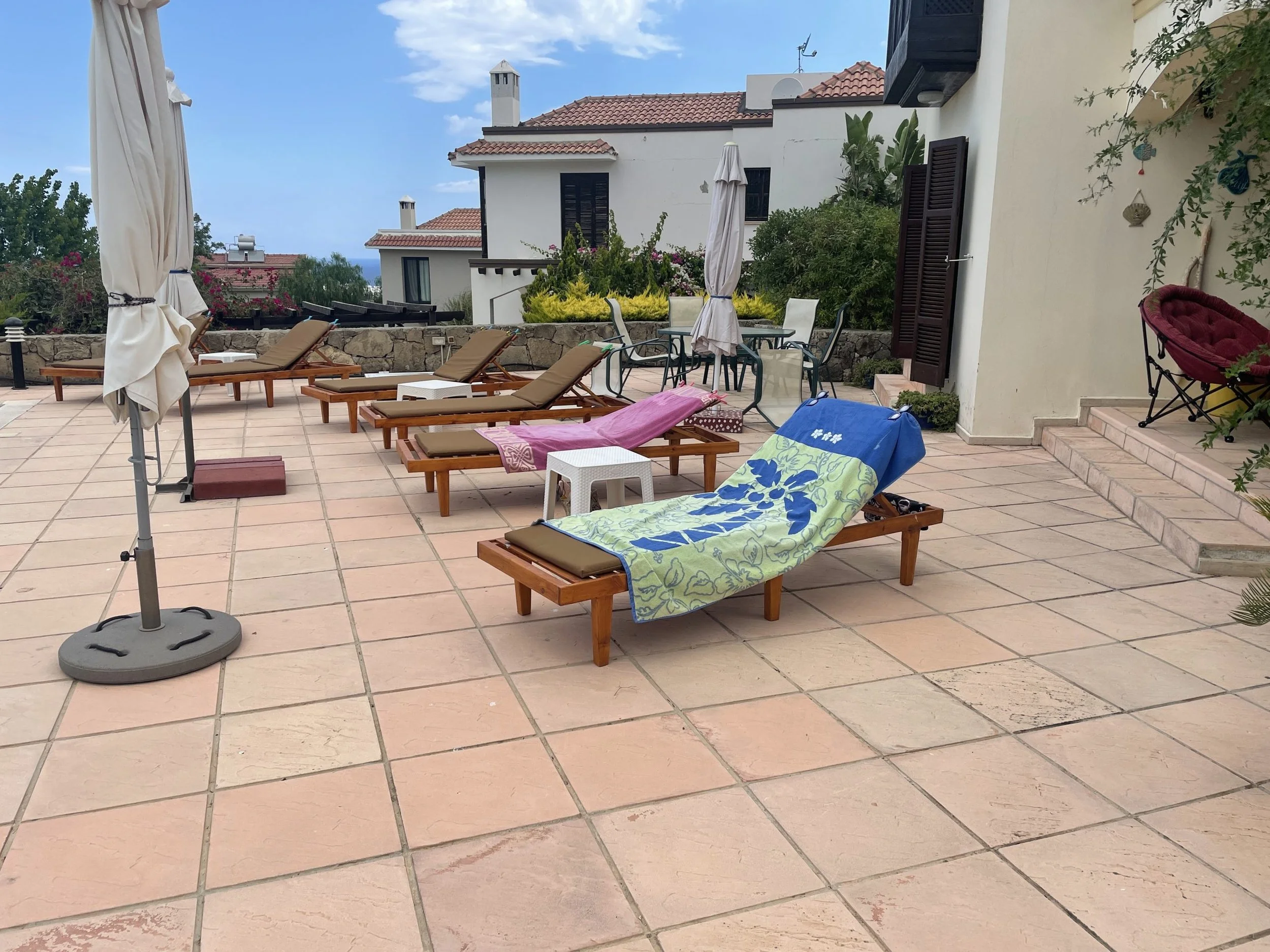 An outdoor patio with several lounge chairs, some covered with towels, and a table with chairs. There's a closed umbrella and a stone wall in the background with lush greenery and buildings with red-tiled roofs.