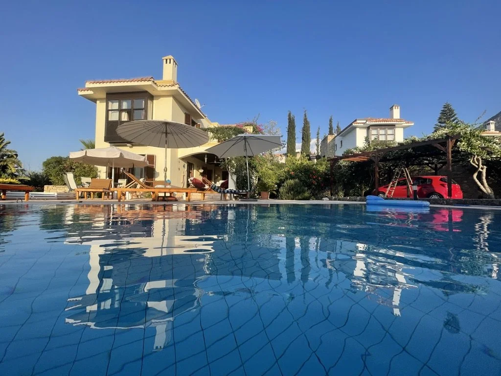 Residential backyard with a swimming pool, umbrellas, lounge chairs, and a house in the background under a clear blue sky.
