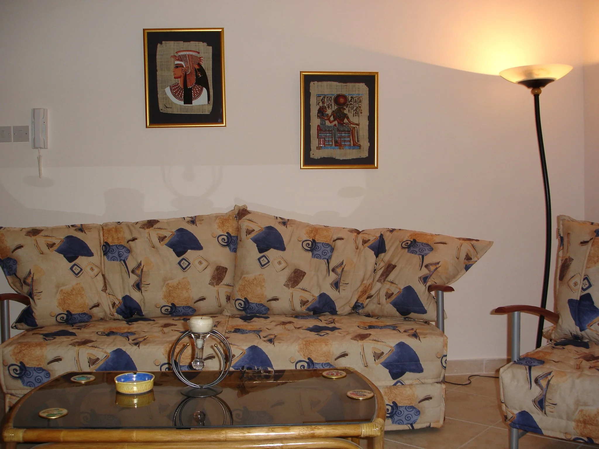 Living room with beige sofa and matching armchair with blue and beige patterned cushions, a glass-topped wooden coffee table with candles, a standing floor lamp, and two framed artworks depicting ancient Egyptian figures on the wall.