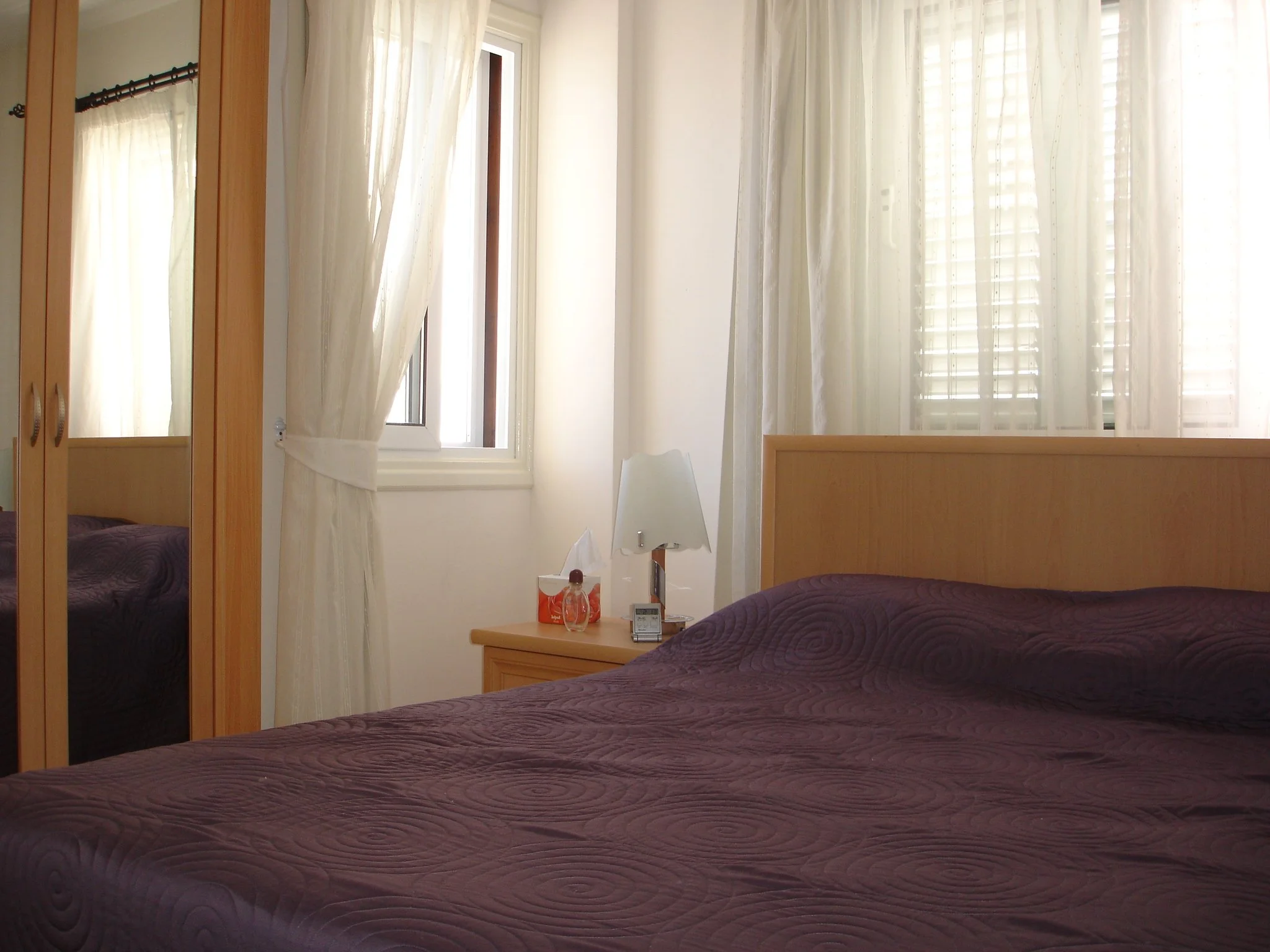 A bedroom with a bed, purple bedspread, wooden headboard, and a nightstand with a lamp, tissue box, and clock, next to a window with sheer curtains.