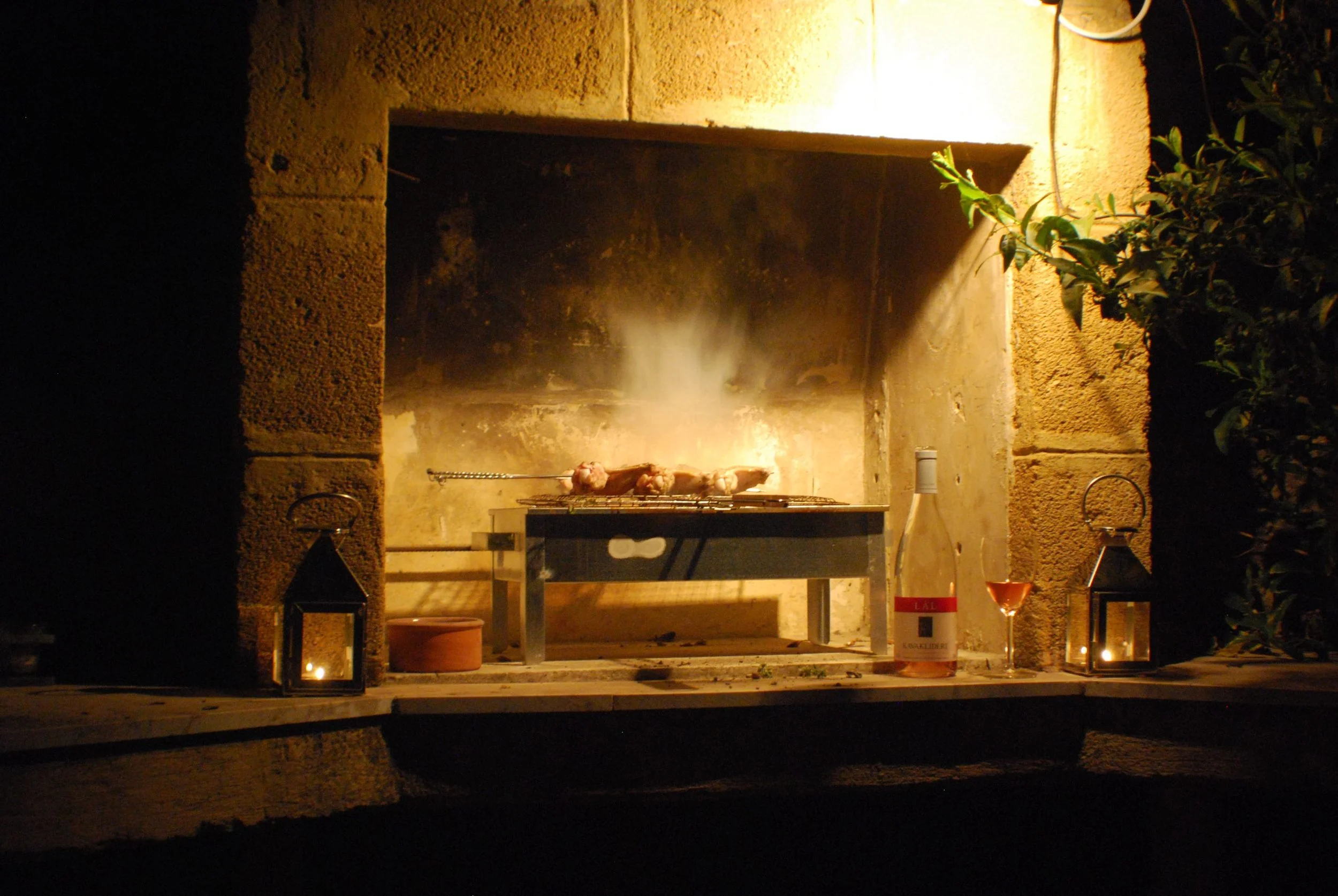 Barbecue grill with skewered meat cooking over an open flame in a cozy outdoor fireplace, surrounded by lanterns and a bottle of rosé wine with a glass, during nighttime.