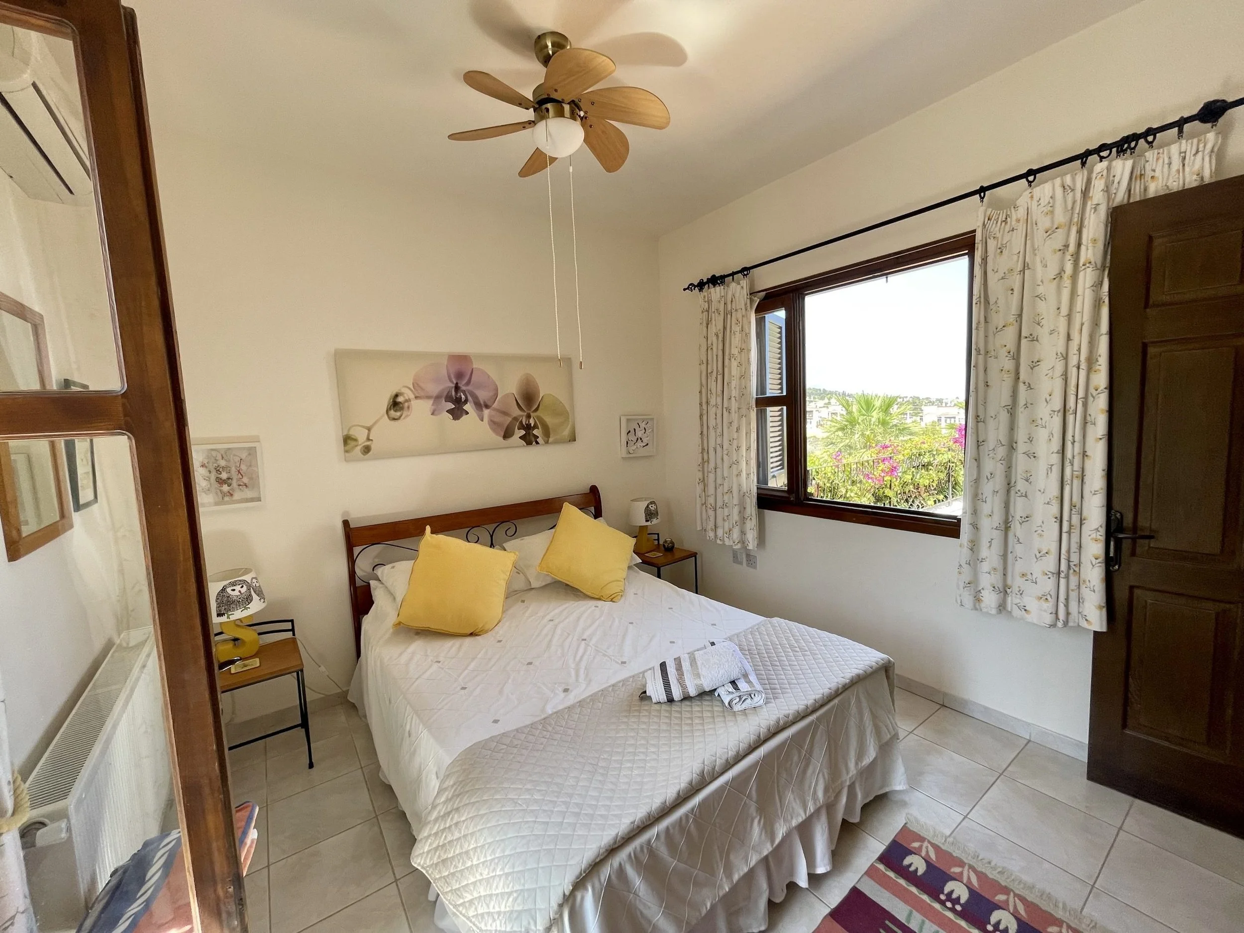 A tidy bedroom with a large bed that has white bedding and two yellow pillows. There are two nightstands next to the bed, each with a small lamp. A window with floral curtains is open, showing greenery outside. A ceiling fan with wooden blades is mou