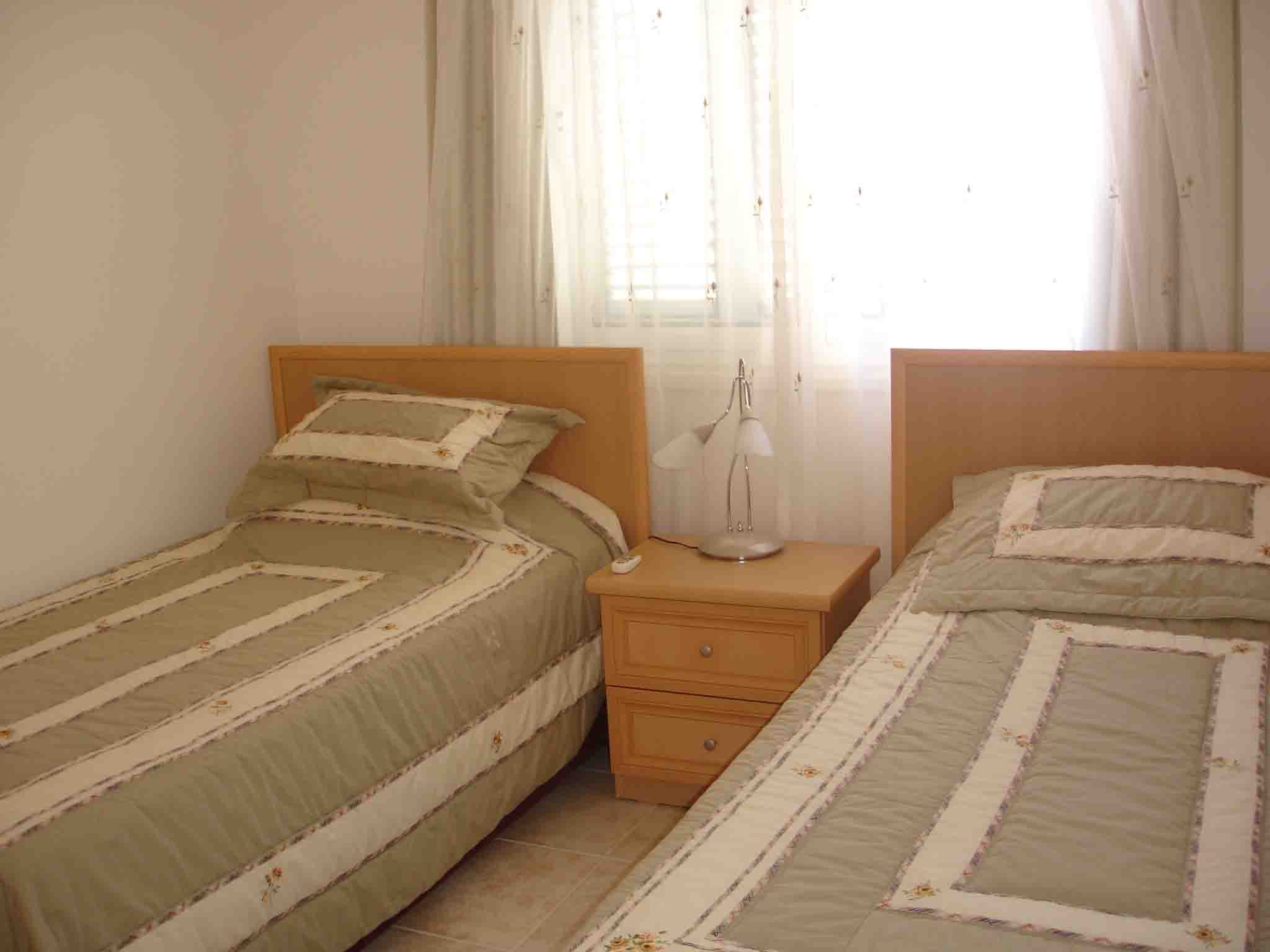 Bedroom with two single beds with beige and green striped bedding. A wooden nightstand with a lamp and a phone sits between the beds. The room has a window with sheer white curtains and a pair of beige curtains.