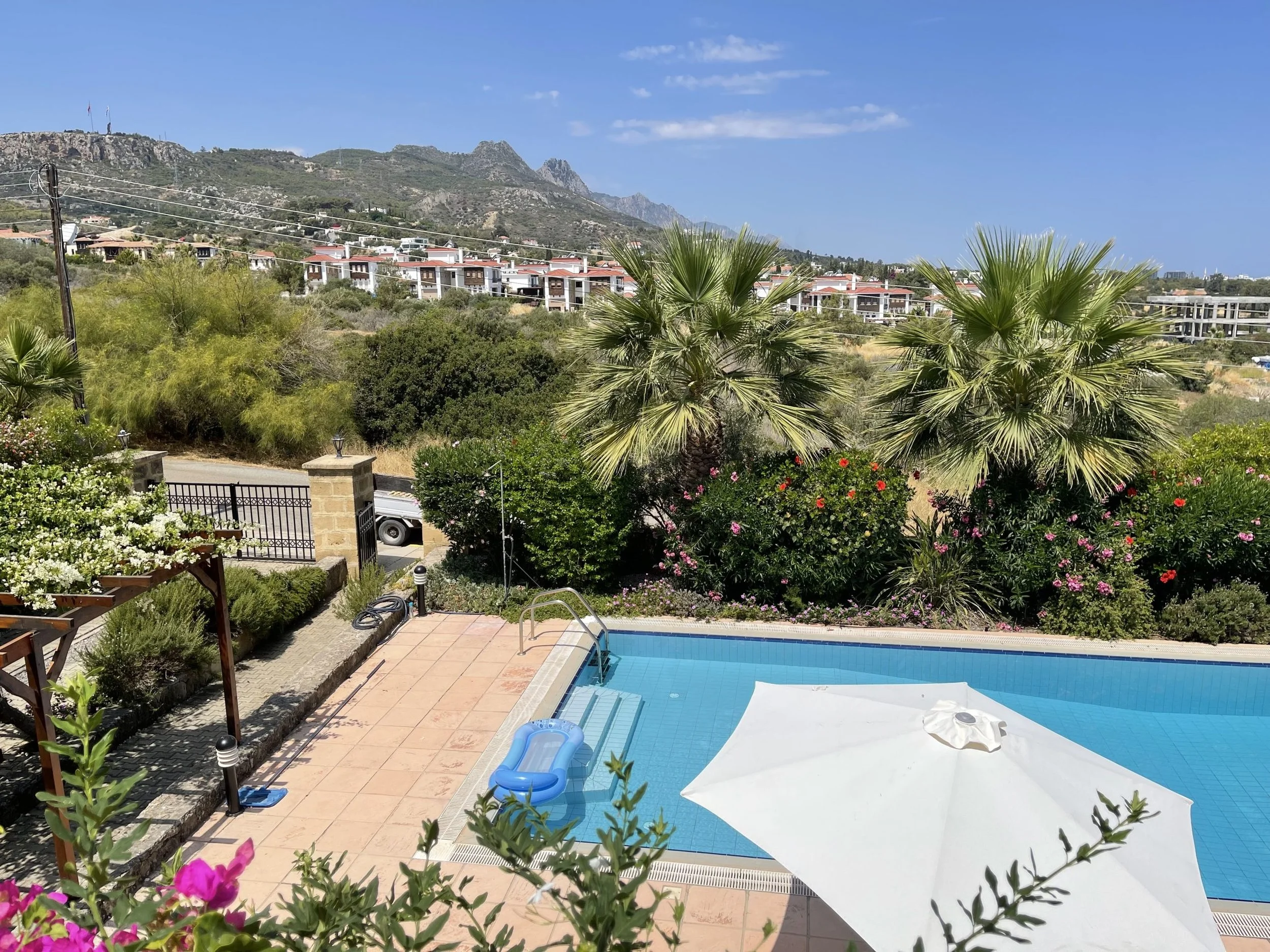 View of a backyard swimming pool with an umbrella, surrounded by bushes and palm trees, overlooking a hilly landscape with houses and mountains in the background.