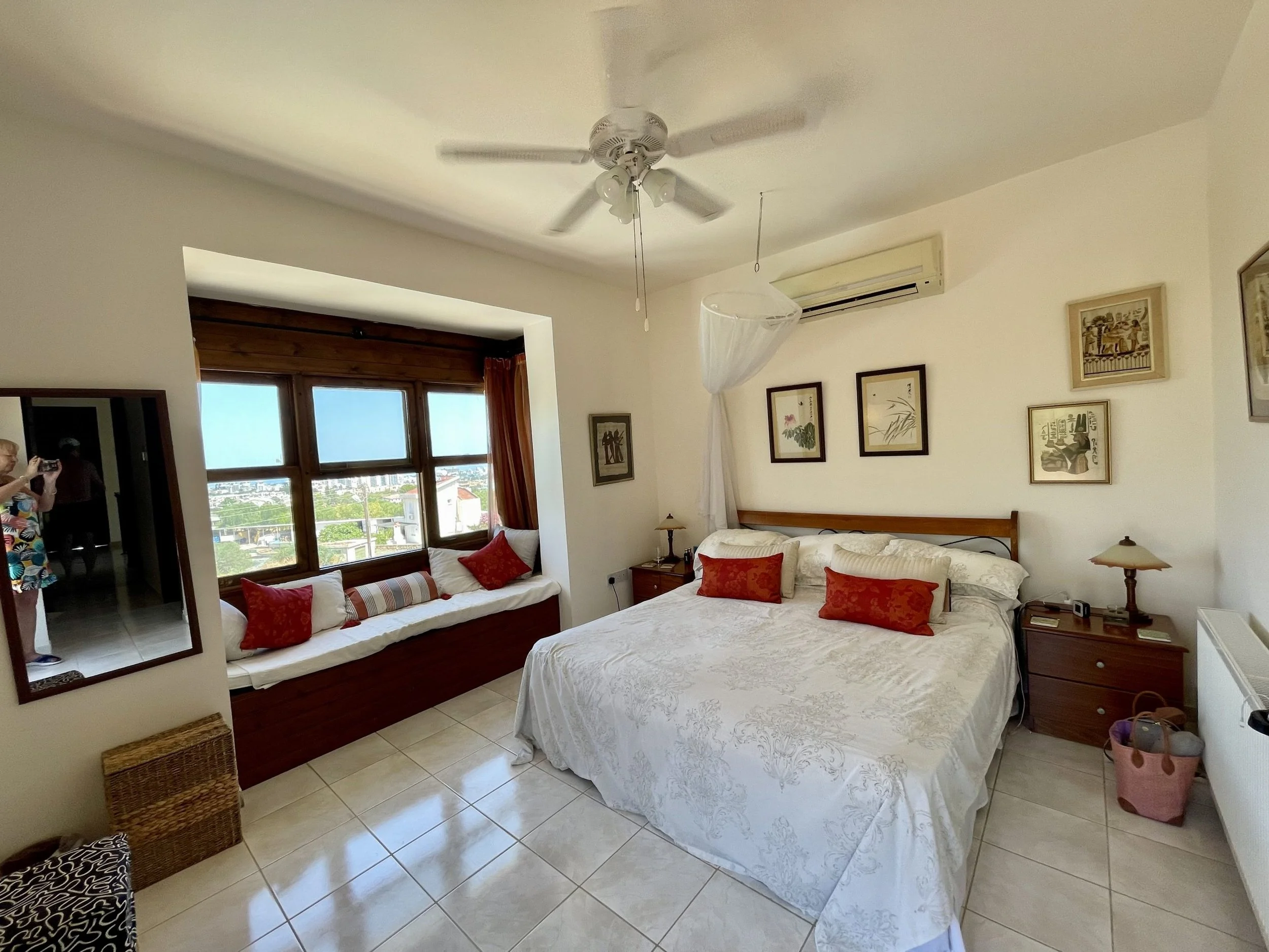 A bedroom with a double bed, nightstands, framed artwork, a built-in window seat with cushions, an air conditioning unit, and ceiling fan.