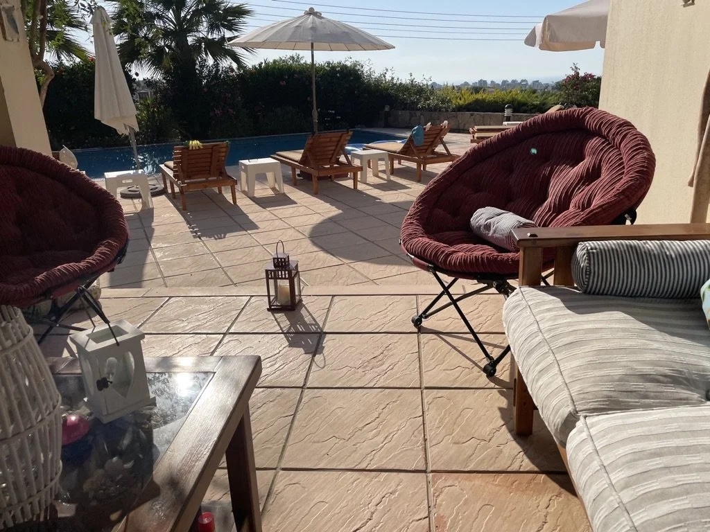 Outdoor patio area with lounge chairs, umbrellas, and a swimming pool in the background, with trees and a clear sky.