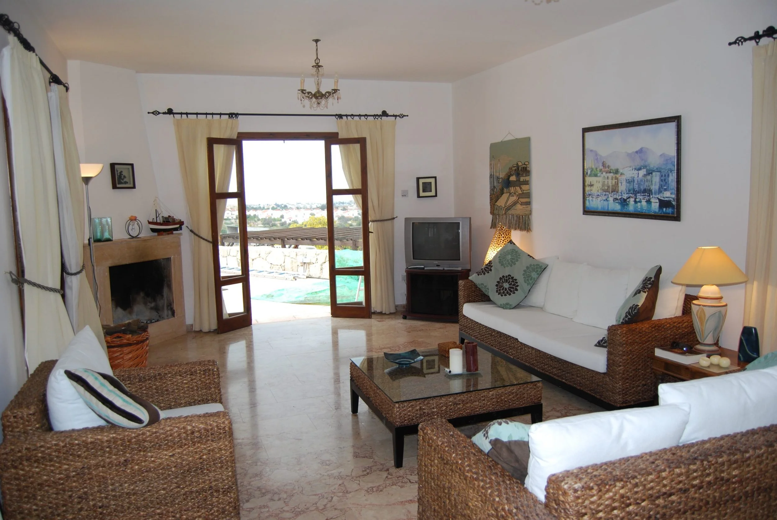 Living room with wicker furniture, white and patterned cushions, a glass-topped coffee table, a fireplace, a small TV, artwork on the walls, and a large open door leading to an outdoor space with a view of a city or town.