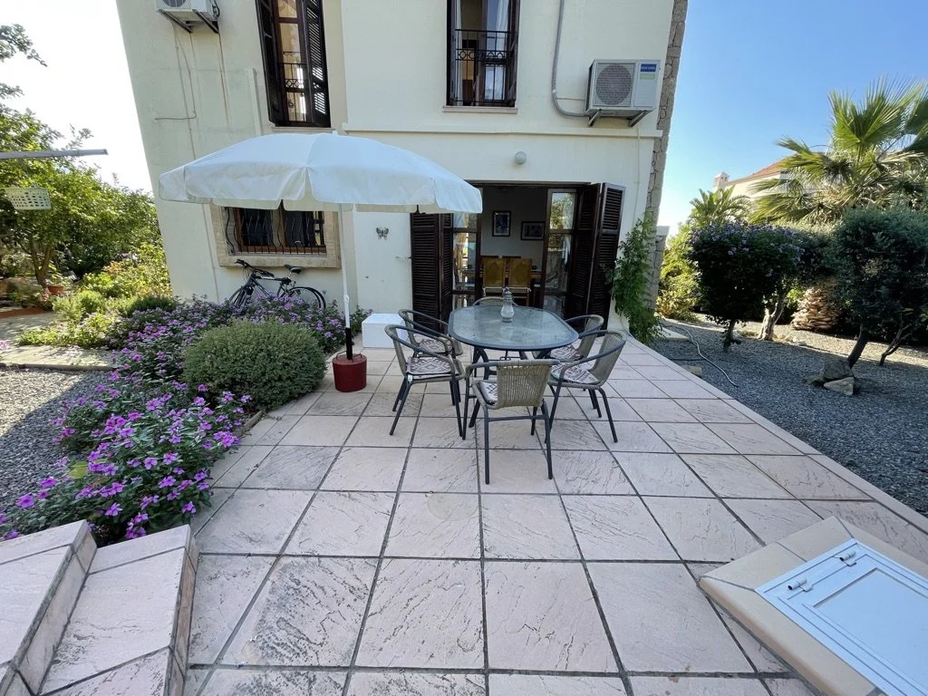 Outdoor patio with a round glass table, six chairs, a white umbrella, and potted plants with purple flowers. A bicycle is parked against the house wall with windows and an air conditioner, and there are trees and bushes in the background.