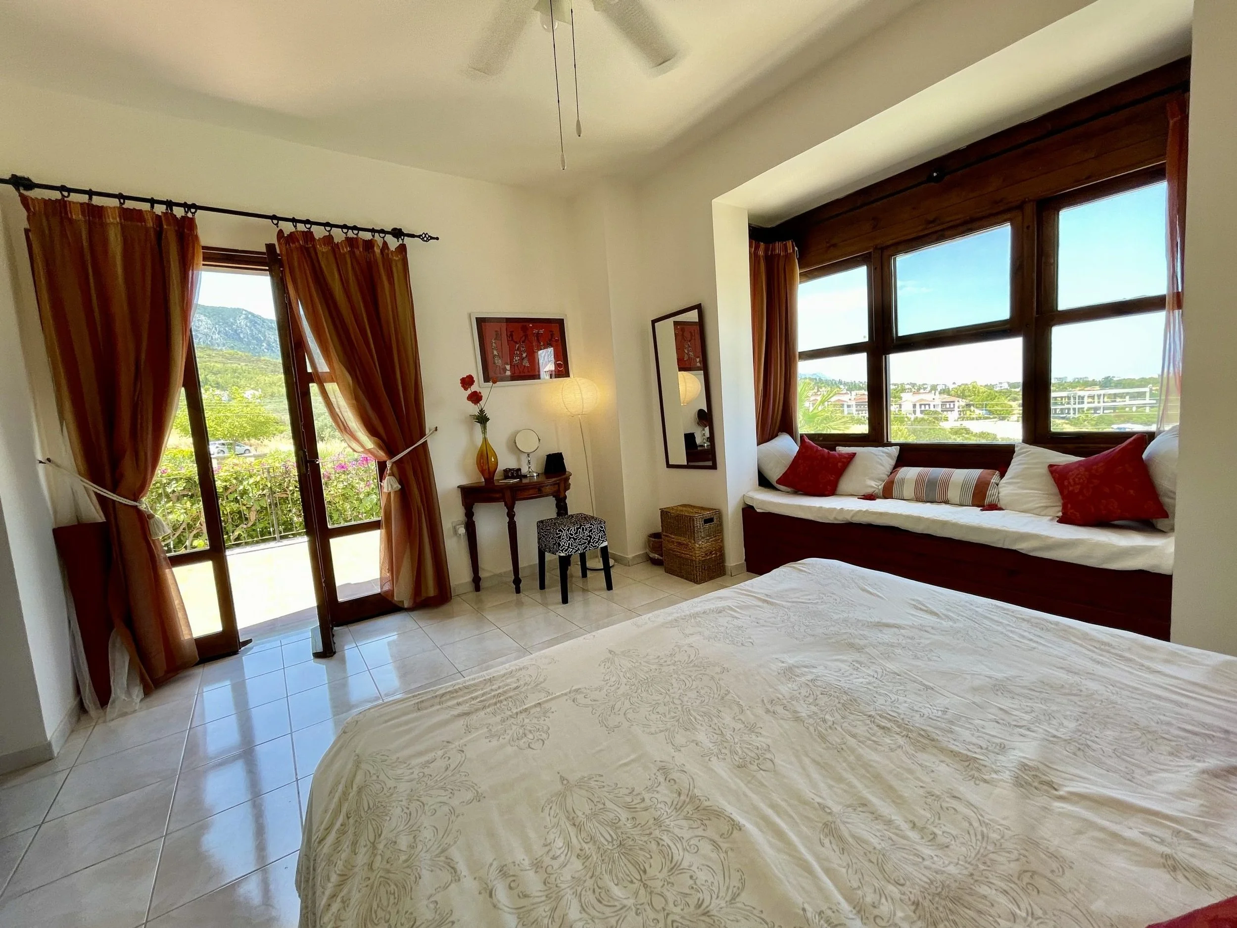 A bright bedroom with a large bed, window seat with pillows, and a balcony door with curtains. Decor includes a small table with a lamp, a mirror, and framed wall art. Outside view shows greenery and buildings.