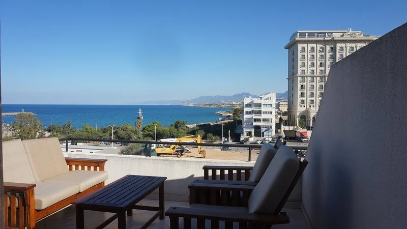 Balcony with outdoor furniture overlooking a beach and ocean, with buildings and a lighthouse in the background.