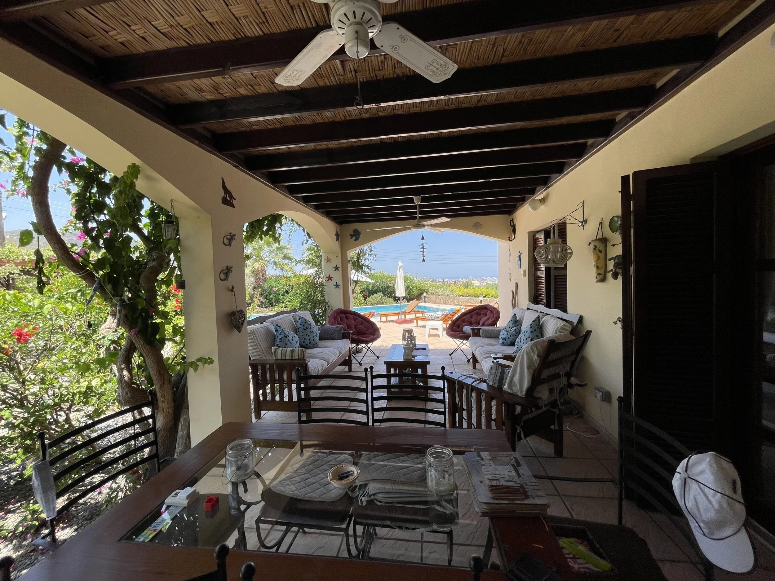 Covered patio area with outdoor seating and view of a pool, lounge chairs, and the ocean in the distance, surrounded by greenery.