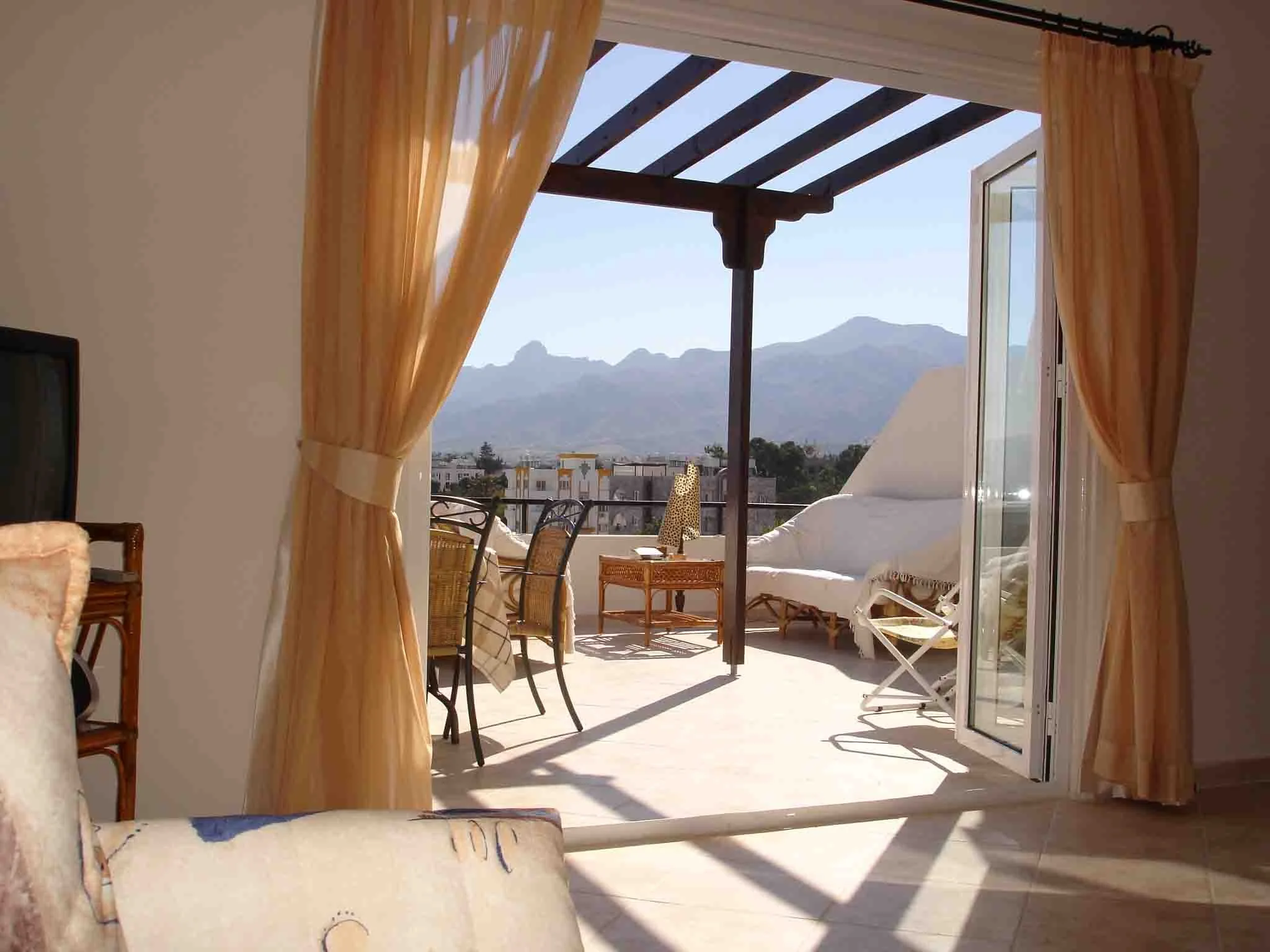 Living room opening to a balcony with mountains in the background, outdoor furniture including a lounge chair and small table, partial view of curtains and furniture inside.