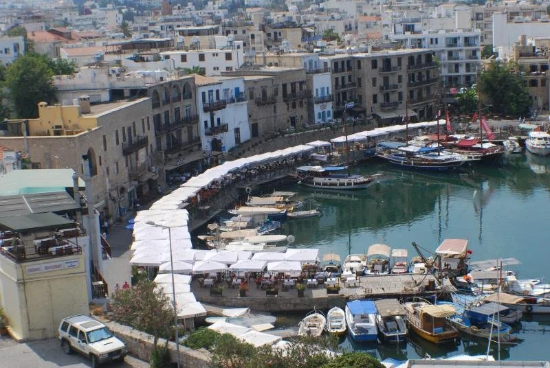 A marina with boats docked, surrounded by white buildings with balconies, and a row of white tents or awnings along the waterfront.