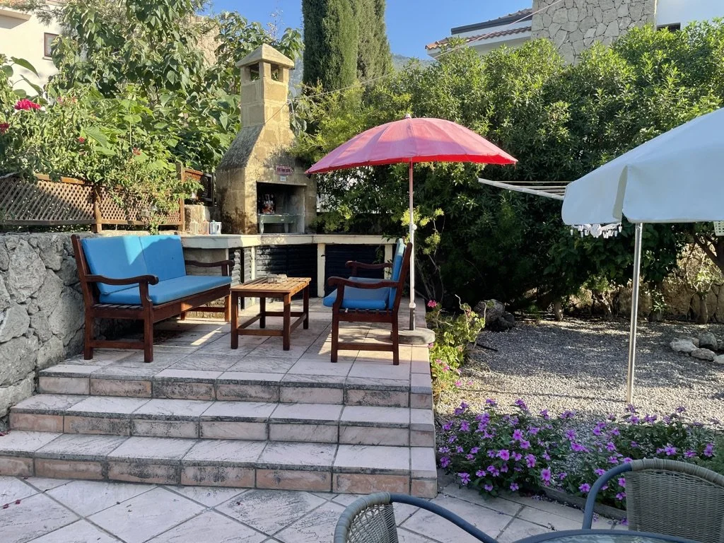 A backyard patio with outdoor furniture, including a blue cushioned bench and chairs, a small wooden table, a red umbrella, and a white umbrella. There is a stone fireplace, lush green bushes, flowers, and a gravel area.