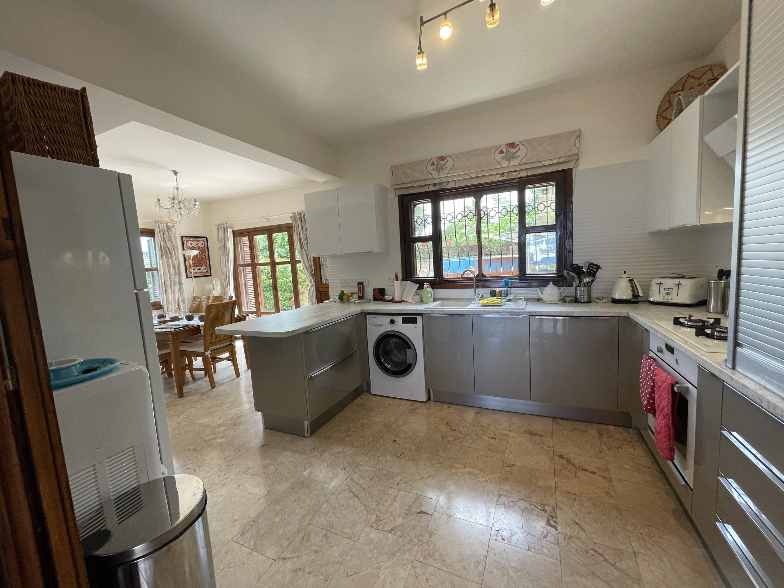 Kitchen with grey cabinets, a window with a floral valance, and common kitchen appliances including a washing machine, oven, toaster, kettle, and coffee maker. A dining area with a chandelier and wooden dining table is visible in the background.