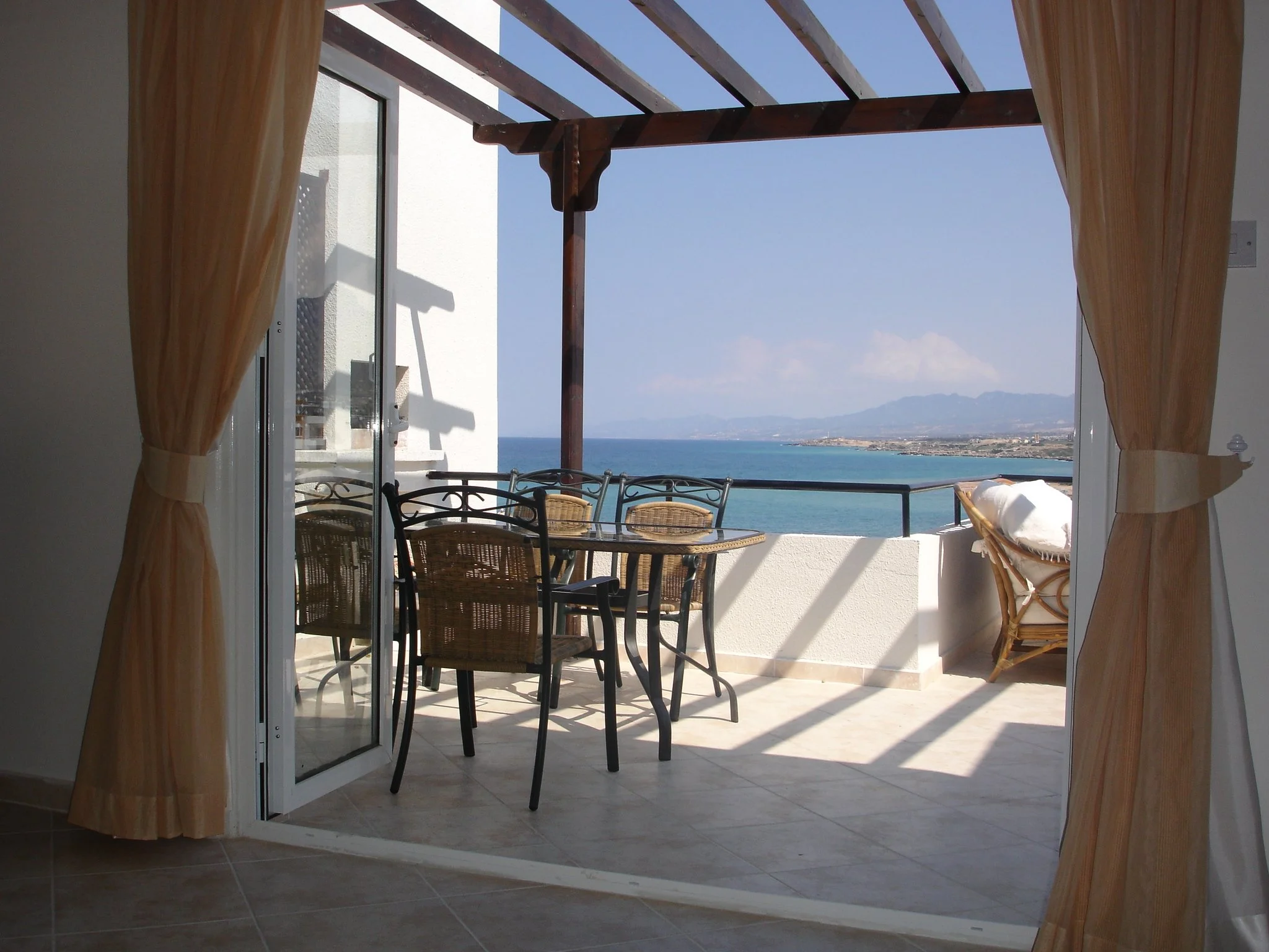 View of a balcony with a table and chairs overlooking the ocean, framed by open sliding doors and curtains, with distant mountains under a blue sky.