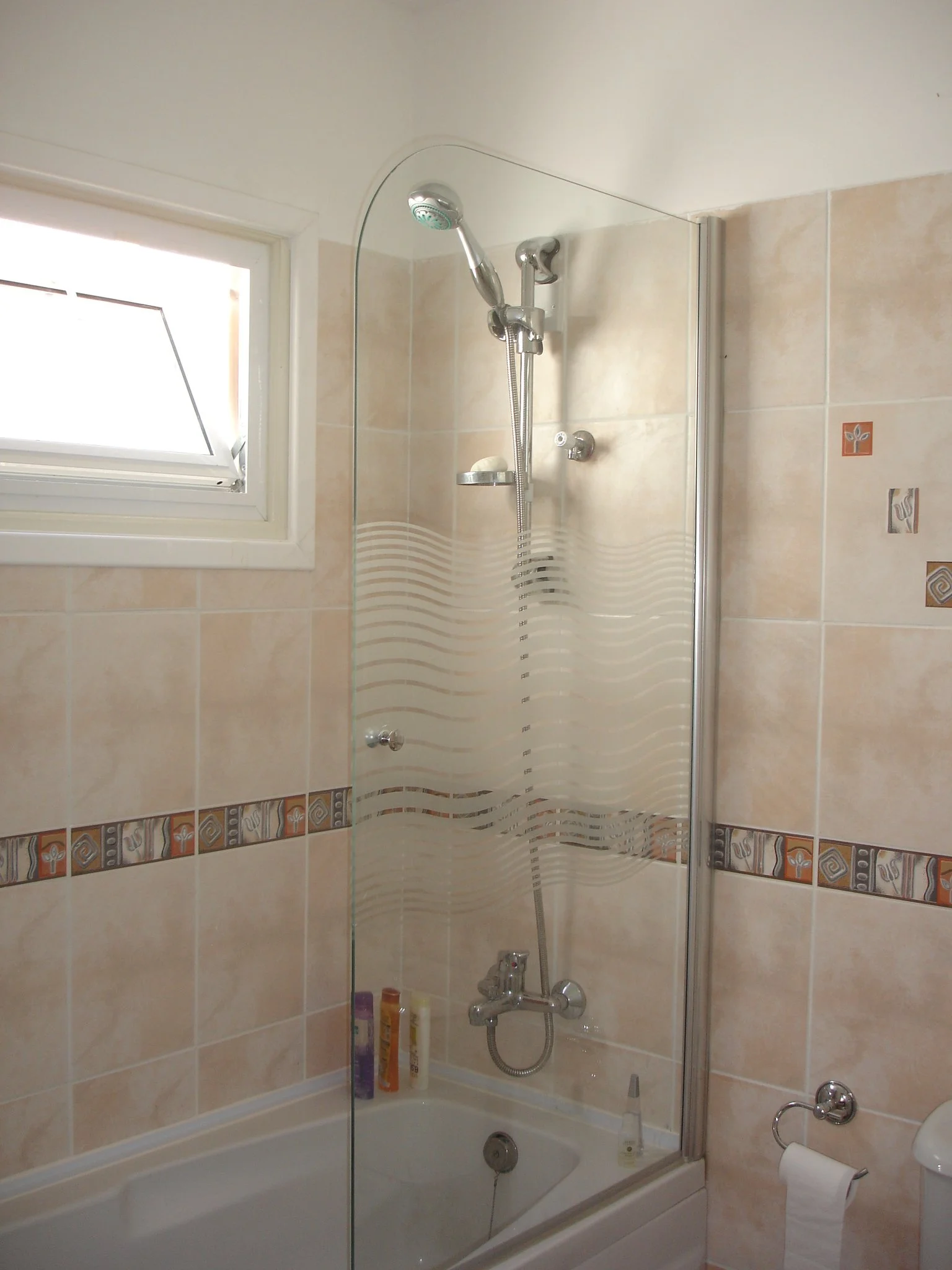 Bathroom with bathtub, glass shower screen, beige tiled walls with decorative border, small window, and toiletries on the tub edge.