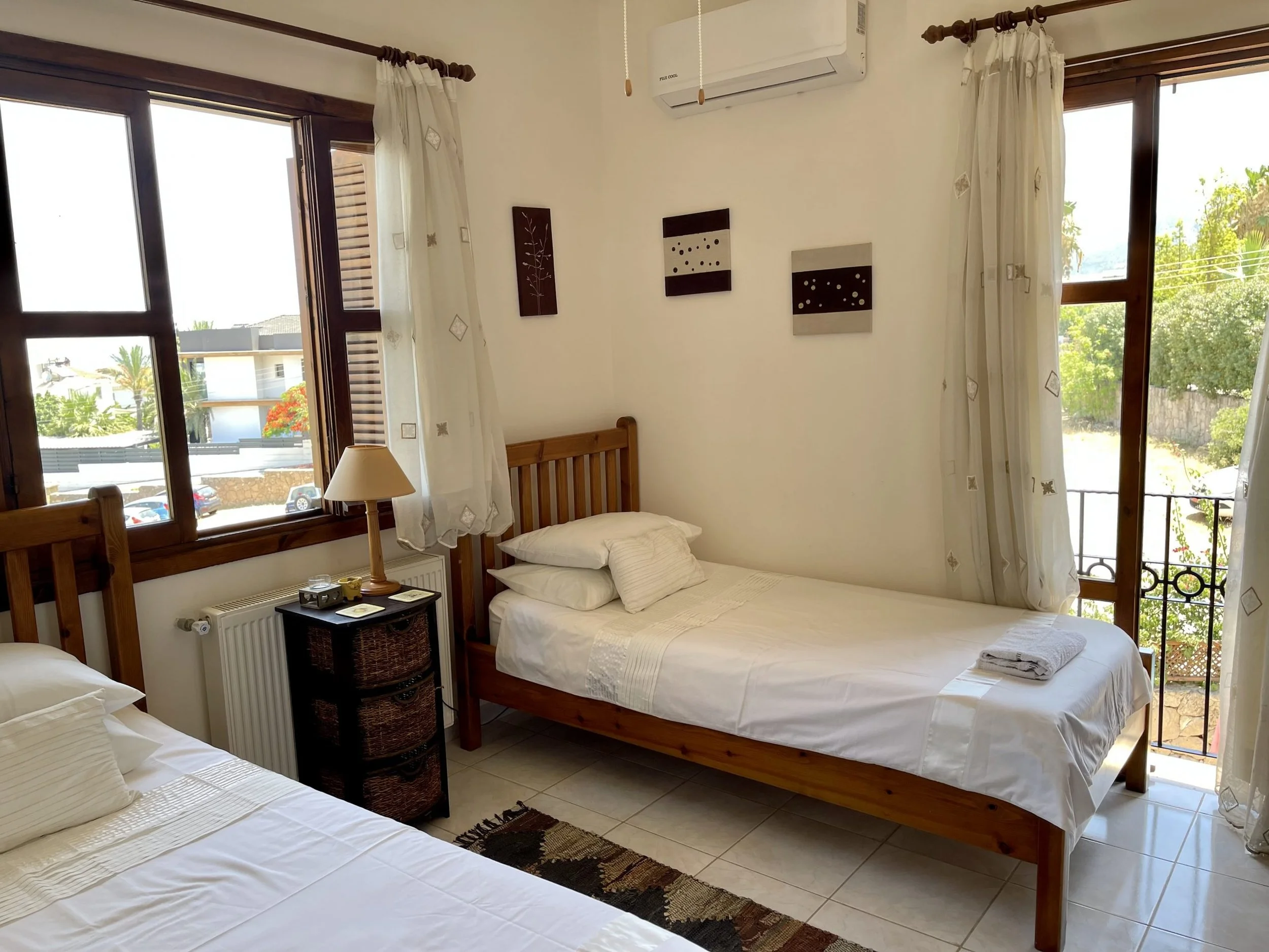 A cozy bedroom with two wooden beds, a window and balcony door with white curtains, a nightstand with a lamp, and decorative wall art.
