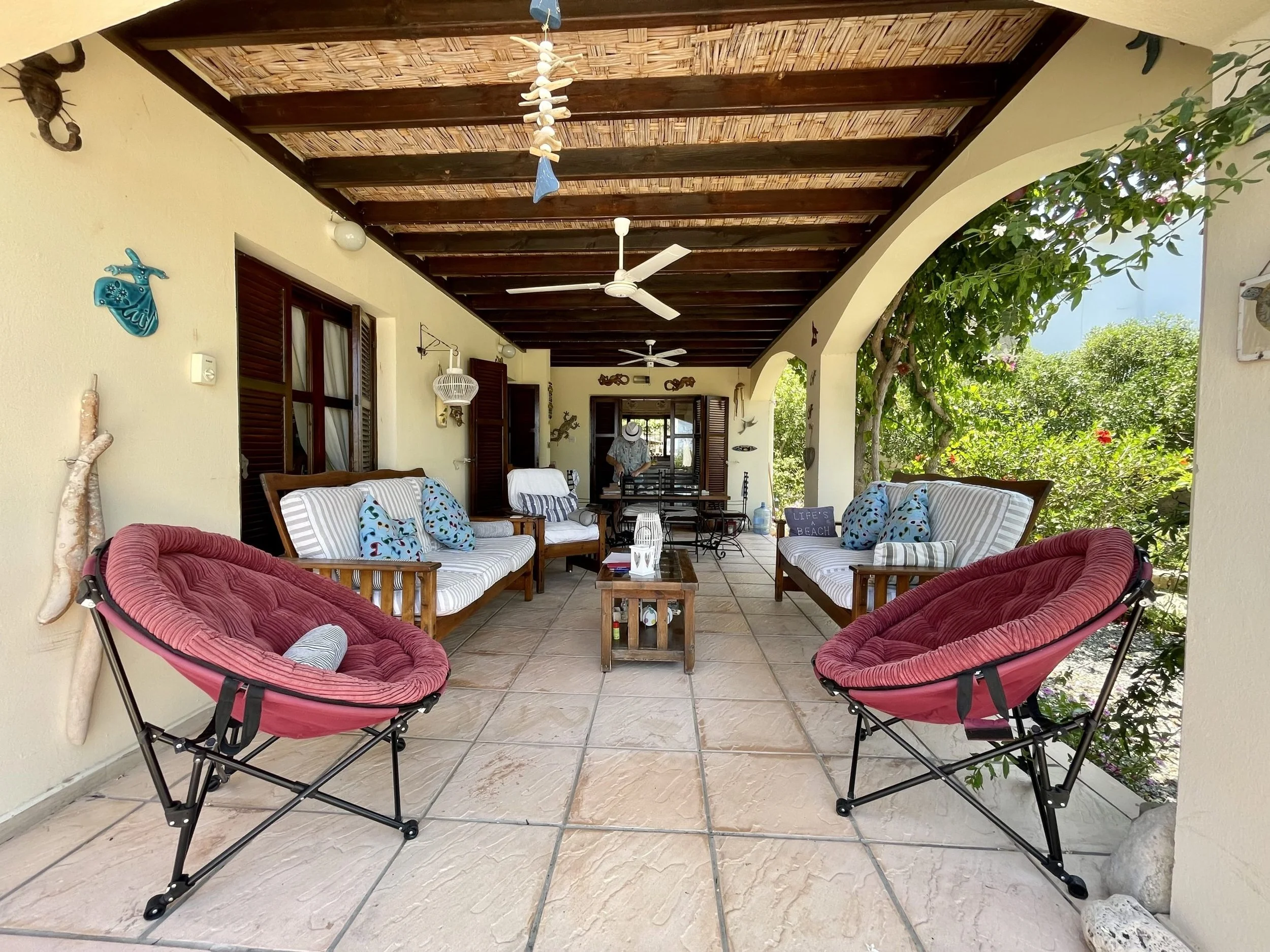 Covered patio with two pink cushioned hanging chairs, four wooden benches with striped and patterned cushions, a small wooden table, ceiling fans, and decorative wall hangings, overlooking green foliage.