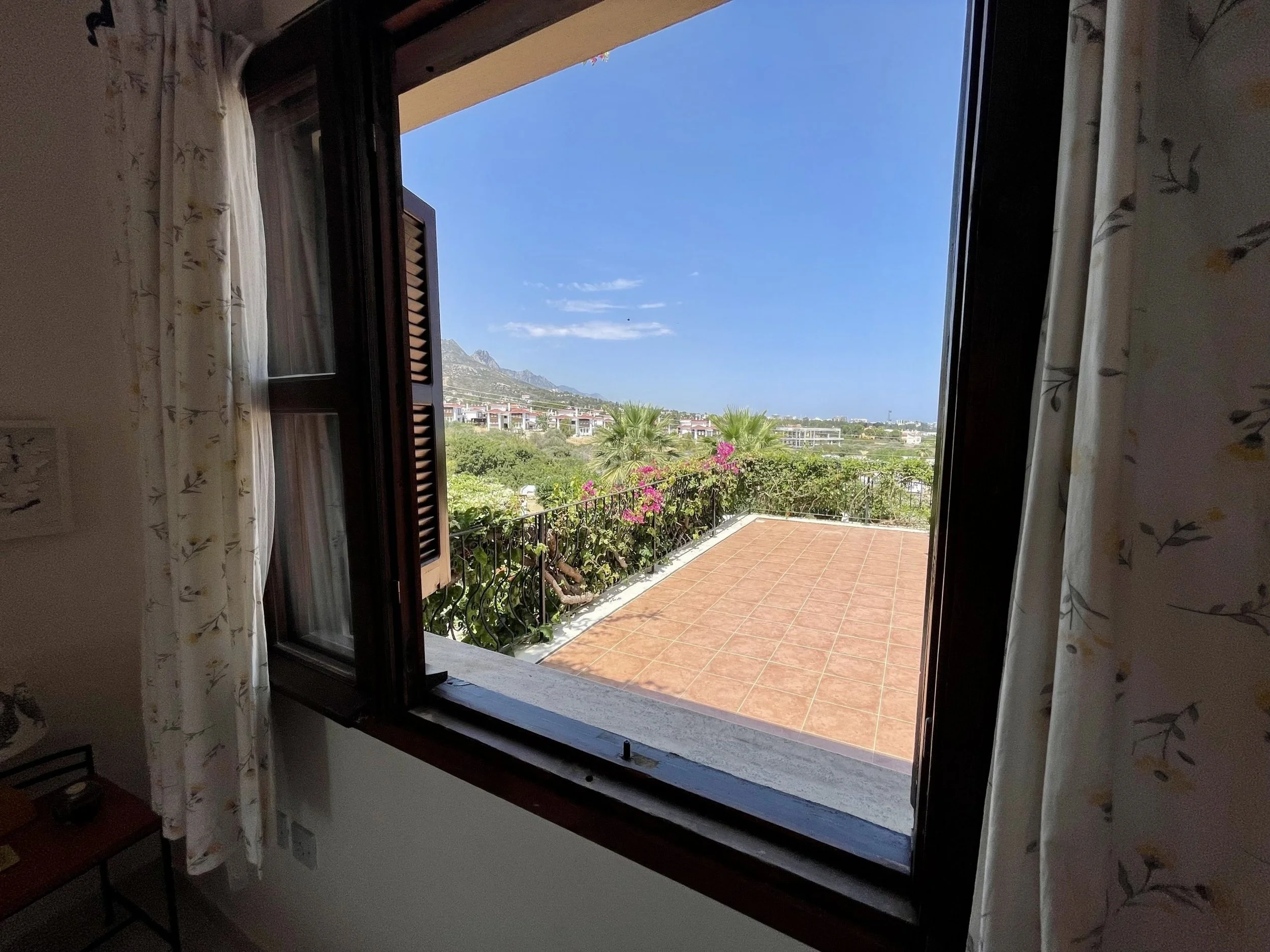 Open window with dark wooden shutters and white floral curtains, revealing a view of a tiled terrace, green bushes, pink flowers, palm trees, and distant mountains under a blue sky.