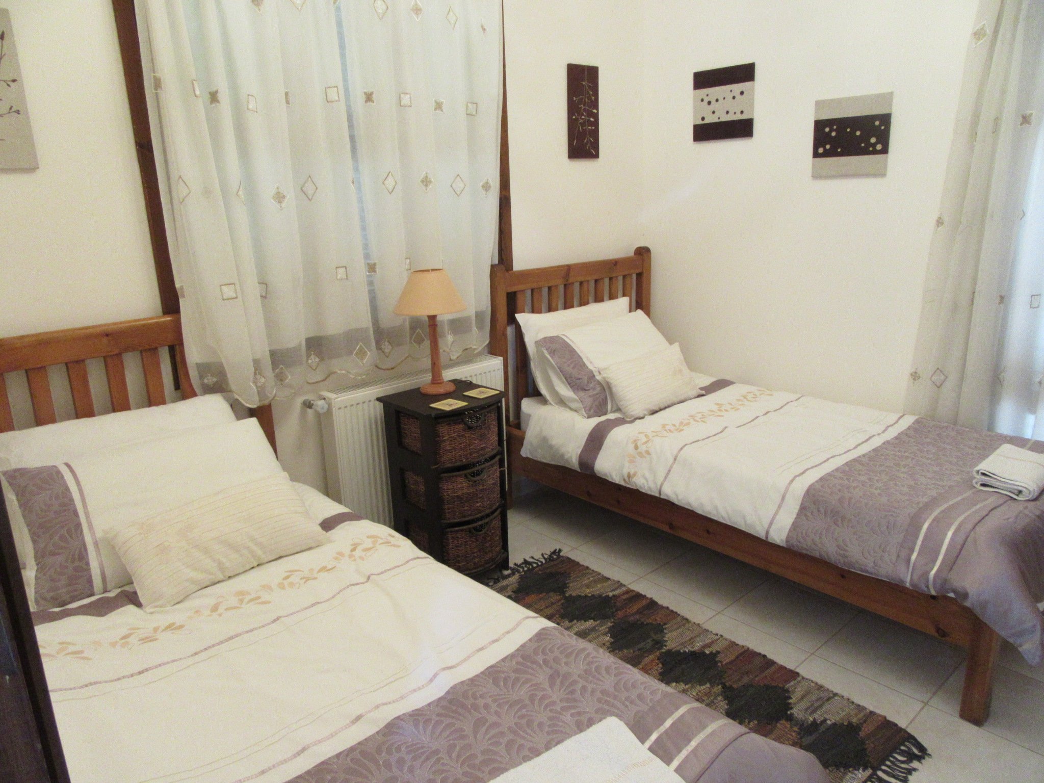 A bedroom with two single beds with wooden headboards and cream-colored bedding, a small dark nightstand with three woven baskets, a beige table lamp, a rug with a geometric pattern, curtains with a diamond pattern, and three black and white abstract