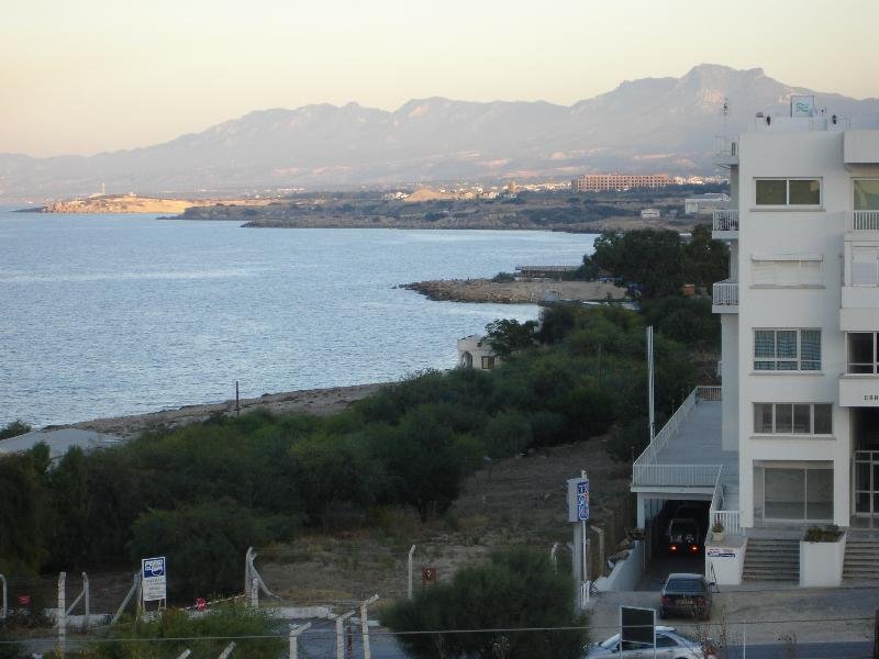A coastal scene with a large body of water, rocky shoreline, green vegetation, and a white apartment building with balconies in the foreground. Mountains are visible in the background.