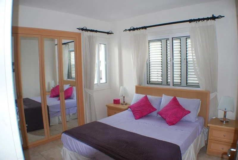 Bedroom with a bed, pink pillows, white curtains, wooden nightstands, and a wardrobe with mirror doors.