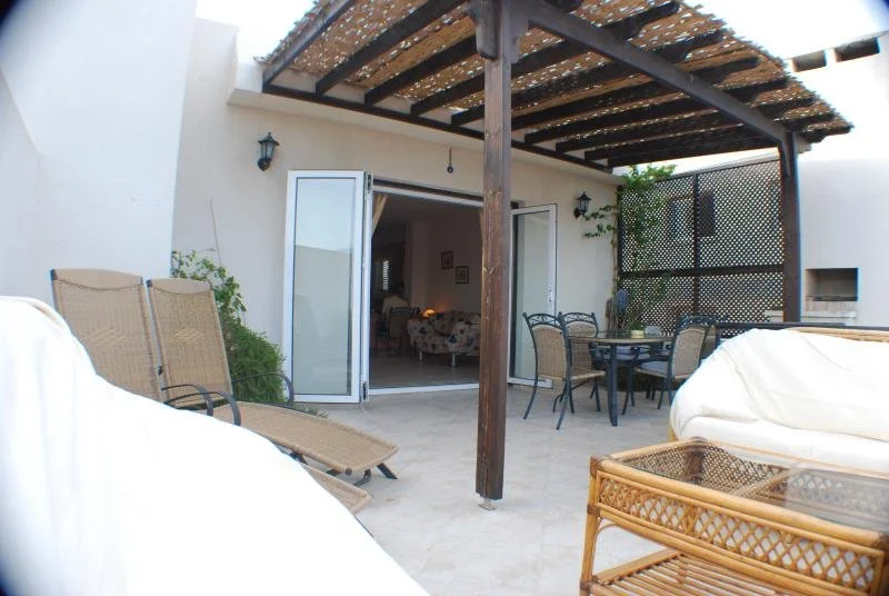 Outdoor patio with wicker chairs, a table, and a white sofa, leading into a room with a floral sofa through open French doors.