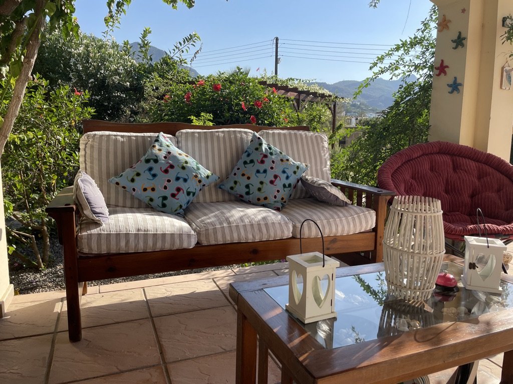 Outdoor patio with a cushioned wooden sofa with striped cushions and colorful pillows, a red cushioned chair, a wooden table with decorative lanterns, surrounded by greenery and mountains in the background.