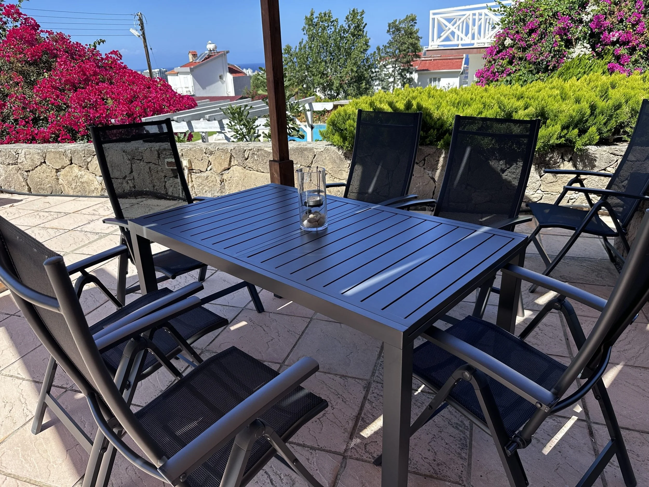 Outdoor patio table with six black chairs, decorative glass vase in the center, surrounded by flowering bushes and houses with a view of the ocean in the background.