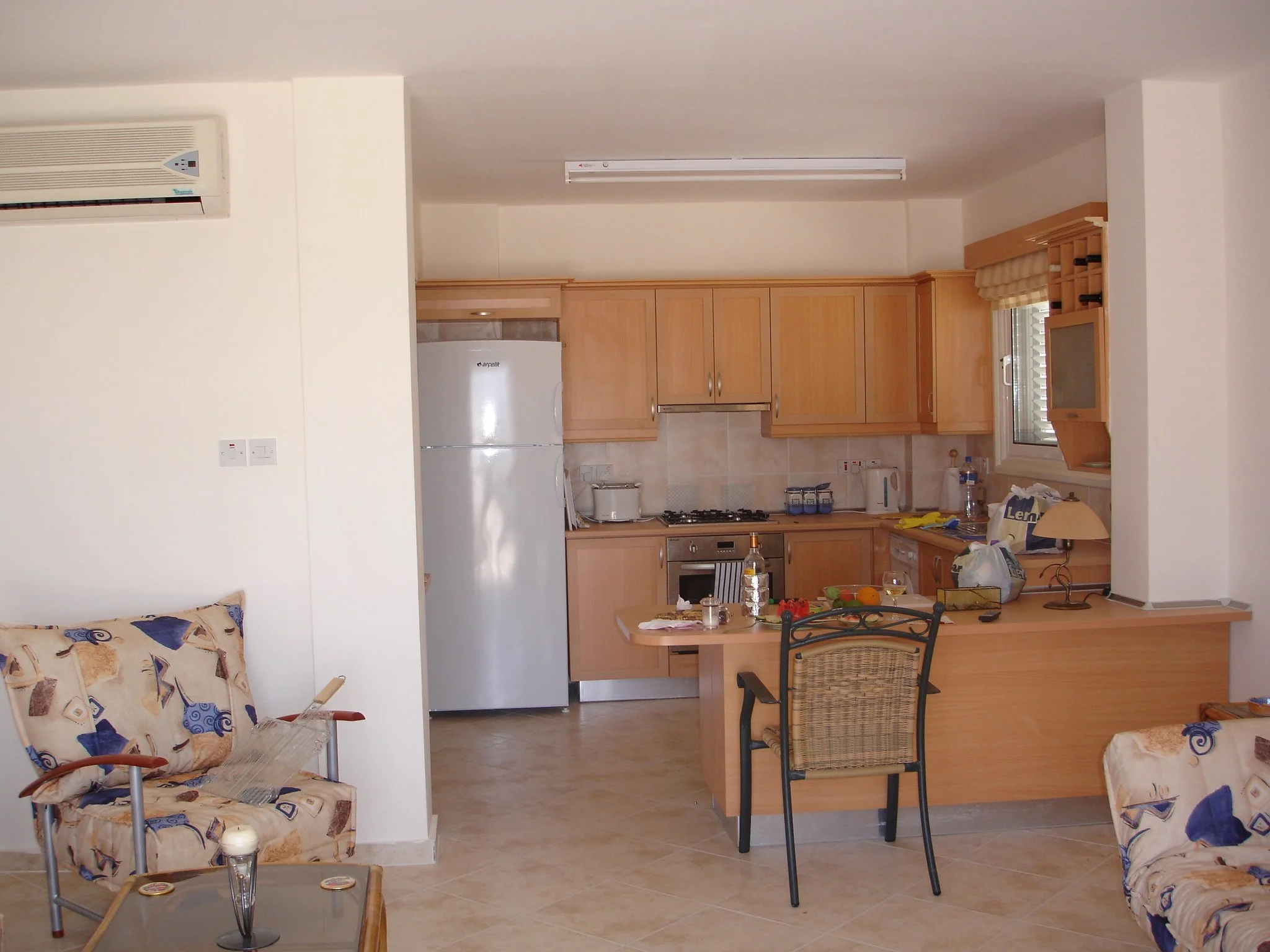 A kitchen with light wooden cabinets, a white refrigerator, a countertop with various kitchen items, and a small dining table with a chair. Two armchairs with patterned fabric are visible in the living area.