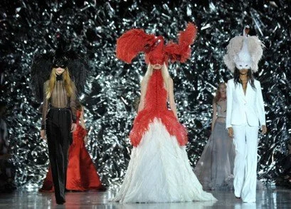 Models walking down a runway during a fashion show, with a silver, sparkly background and elaborate costumes.