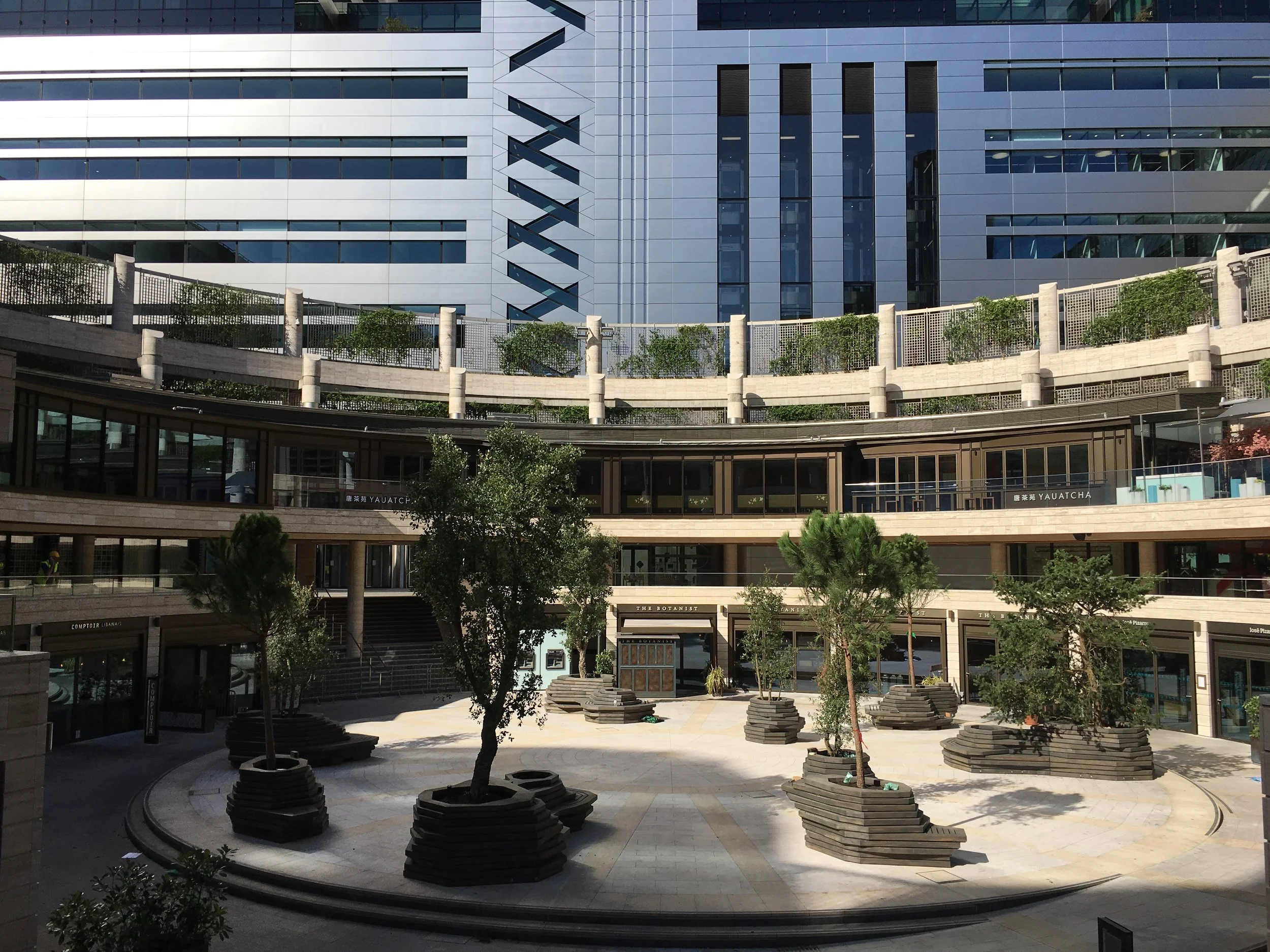 A modern multi-story office building with large glass windows, a staircase with geometric design on the exterior, and a courtyard with trees and circular benches in the foreground.