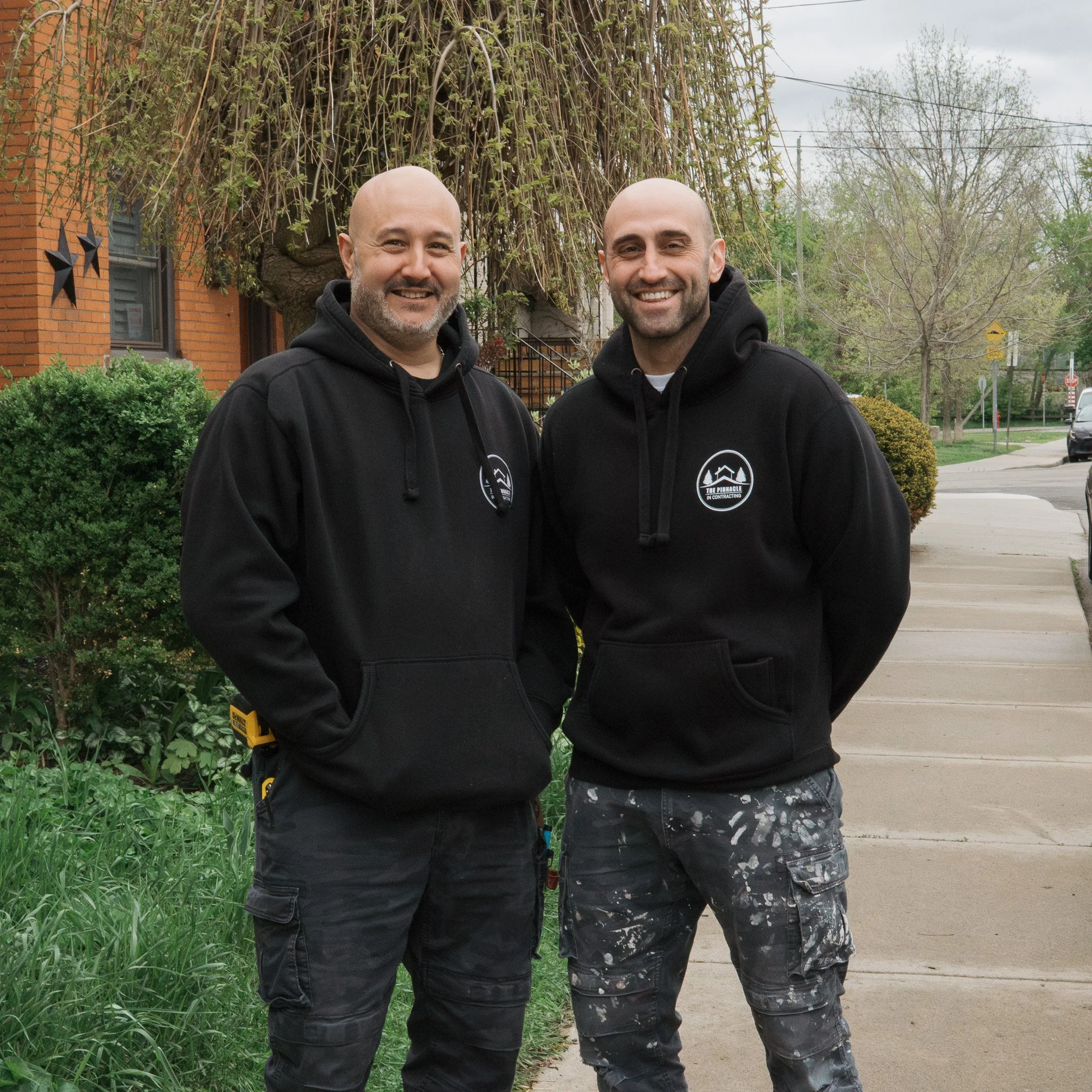 Two men standing outdoors on a sidewalk, smiling at the camera, wearing black hoodies with a logo, with a tree and residential buildings in the background.