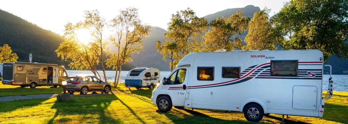 RVs and cars parked by a lake with trees and mountains, during sunset.
