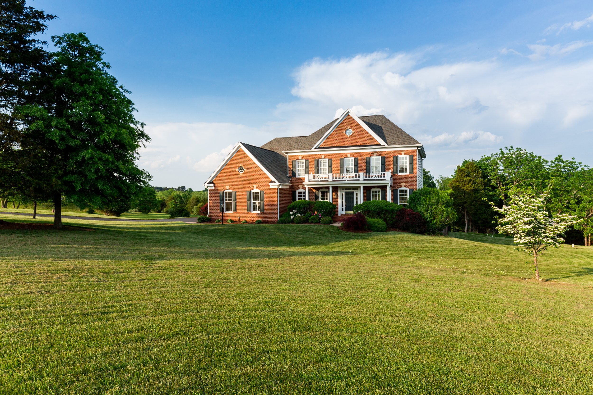 A large brick house with multiple stories, white shutters, and a front porch surrounded by green trees and a well-manicured lawn.