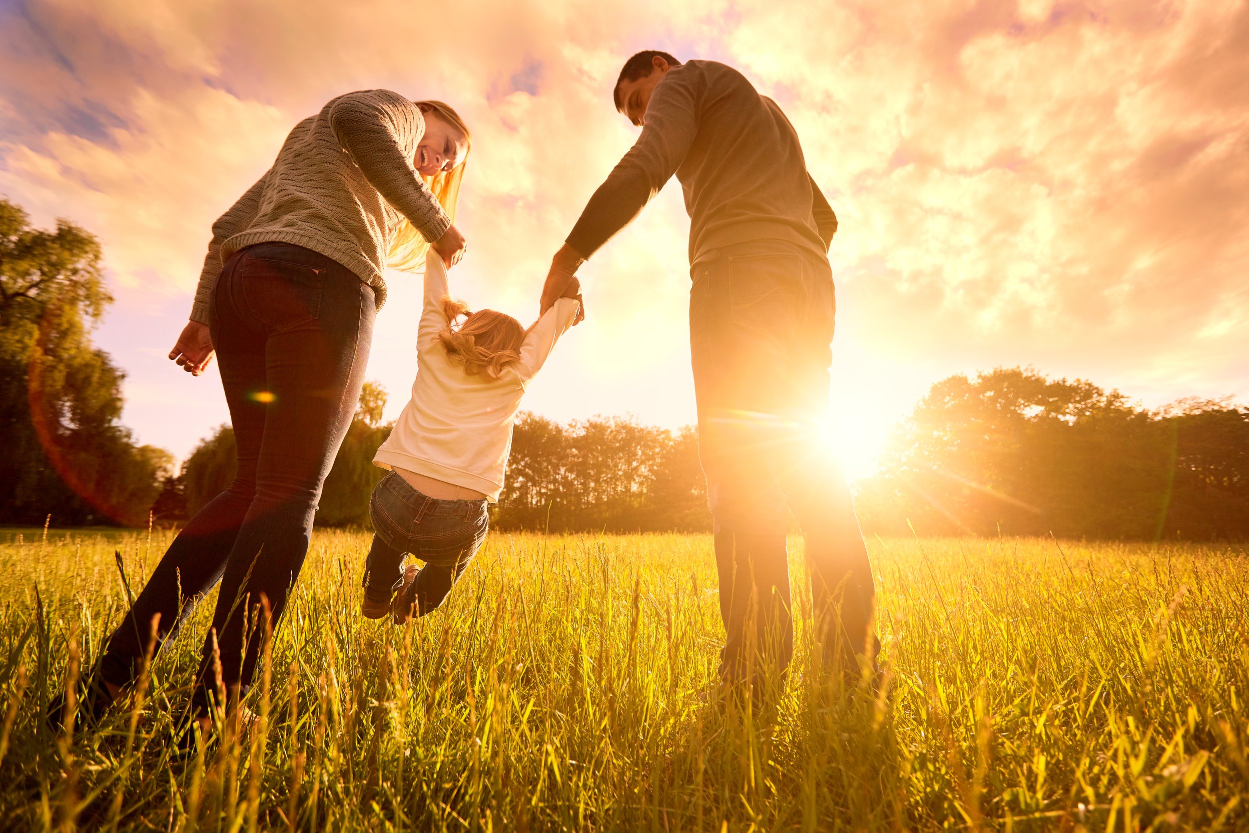 A family of four, including two adults and two children, holding hands and running through a grassy field at sunset. The sky is filled with orange and pink clouds, creating a warm and joyful atmosphere.