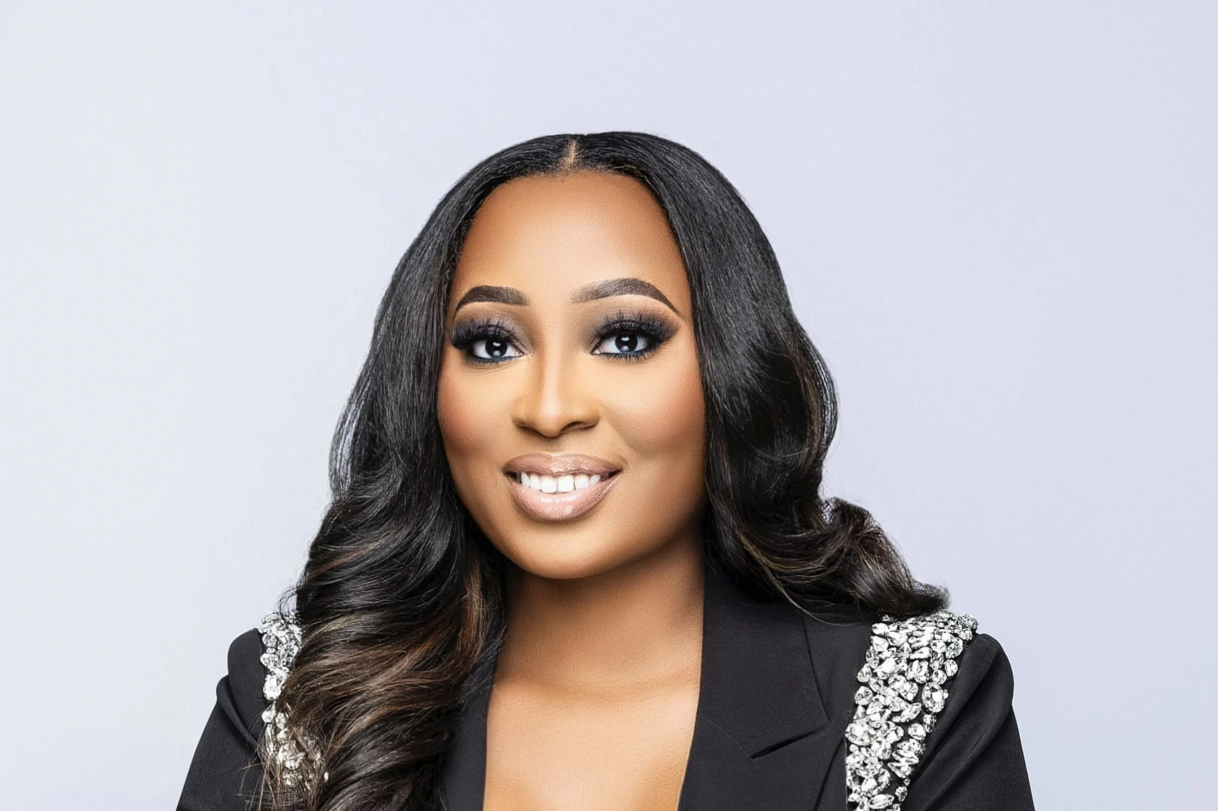A woman with long, wavy black hair styled to the side, wearing makeup with bold eye makeup and nude lipstick, dressed in a black blazer with decorative silver embellishments on the shoulders, smiling against a plain light gray background.