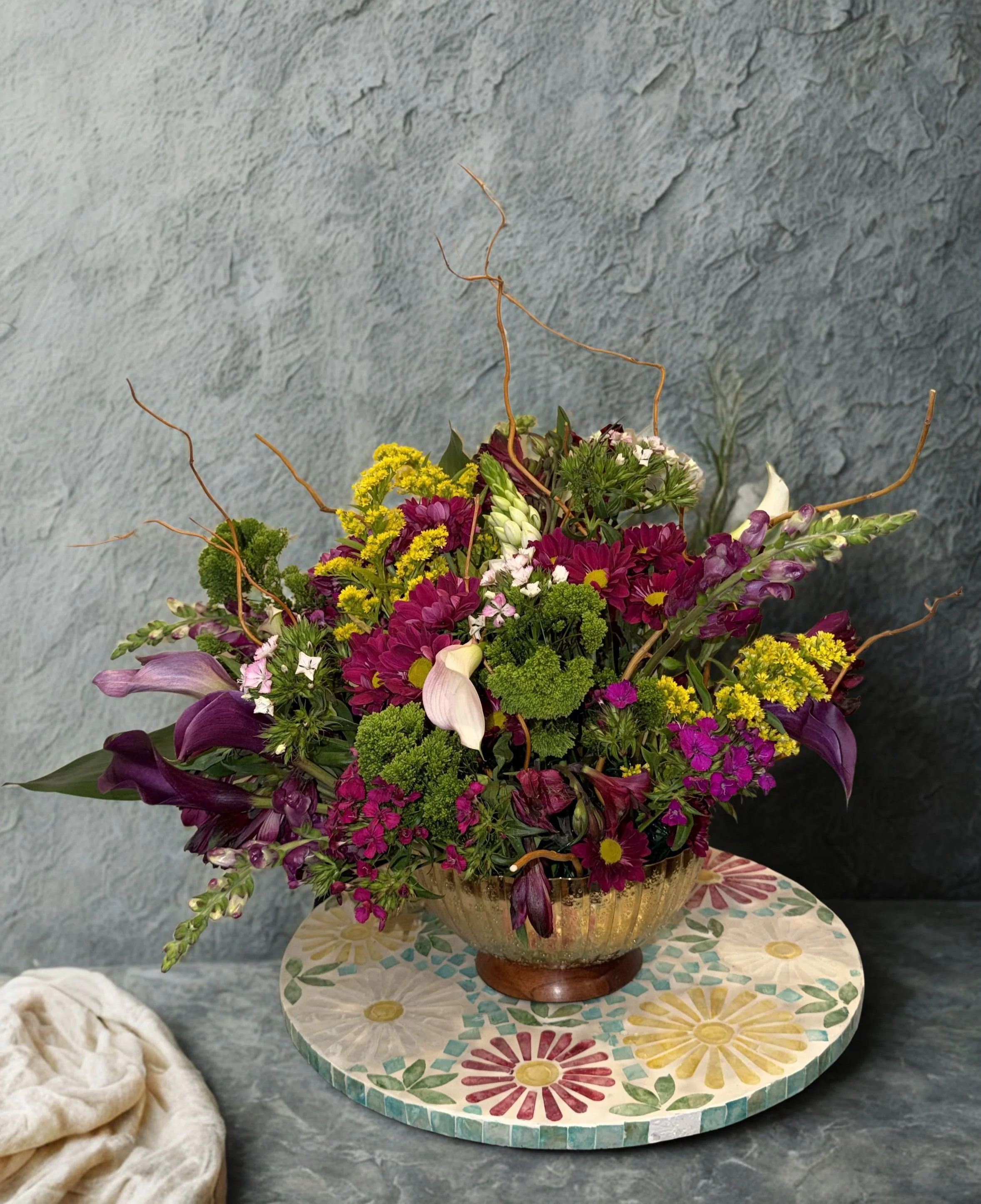 A colorful bouquet of purple, pink, yellow, and white flowers in a decorative gold and bronze vase on a floral-patterned round board.