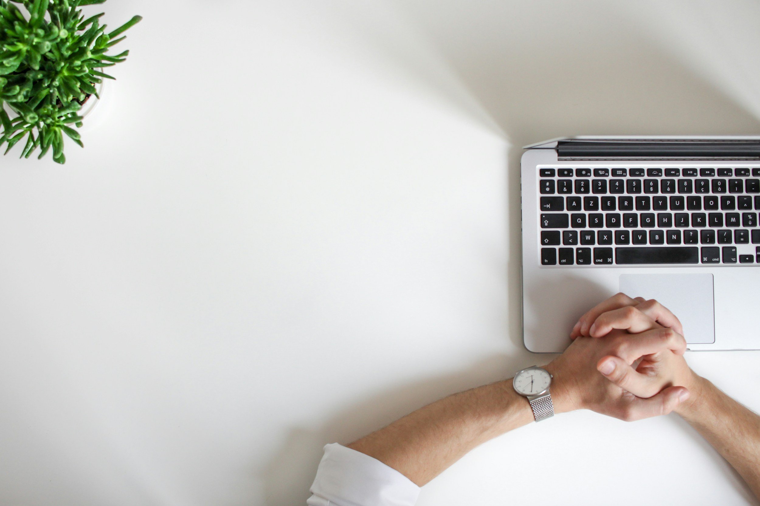 A person with a white shirt and a wristwatch sitting at a white desk with a silver laptop and a small green plant in the top left corner.