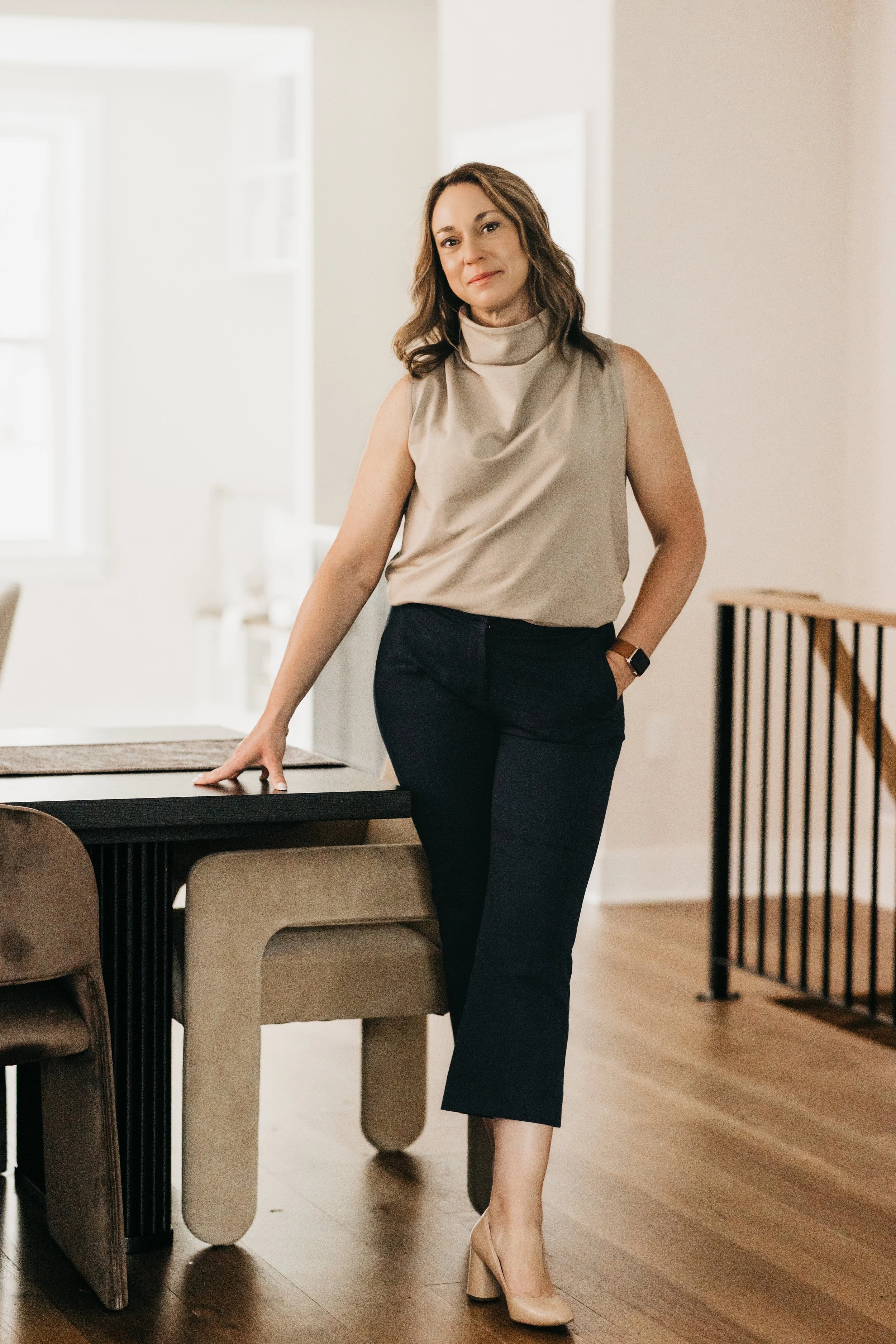 A woman with shoulder-length brown hair, wearing a beige sleeveless top, black ankle-length pants, and beige heels, standing in a well-lit room with wooden floors, leaning on a table.