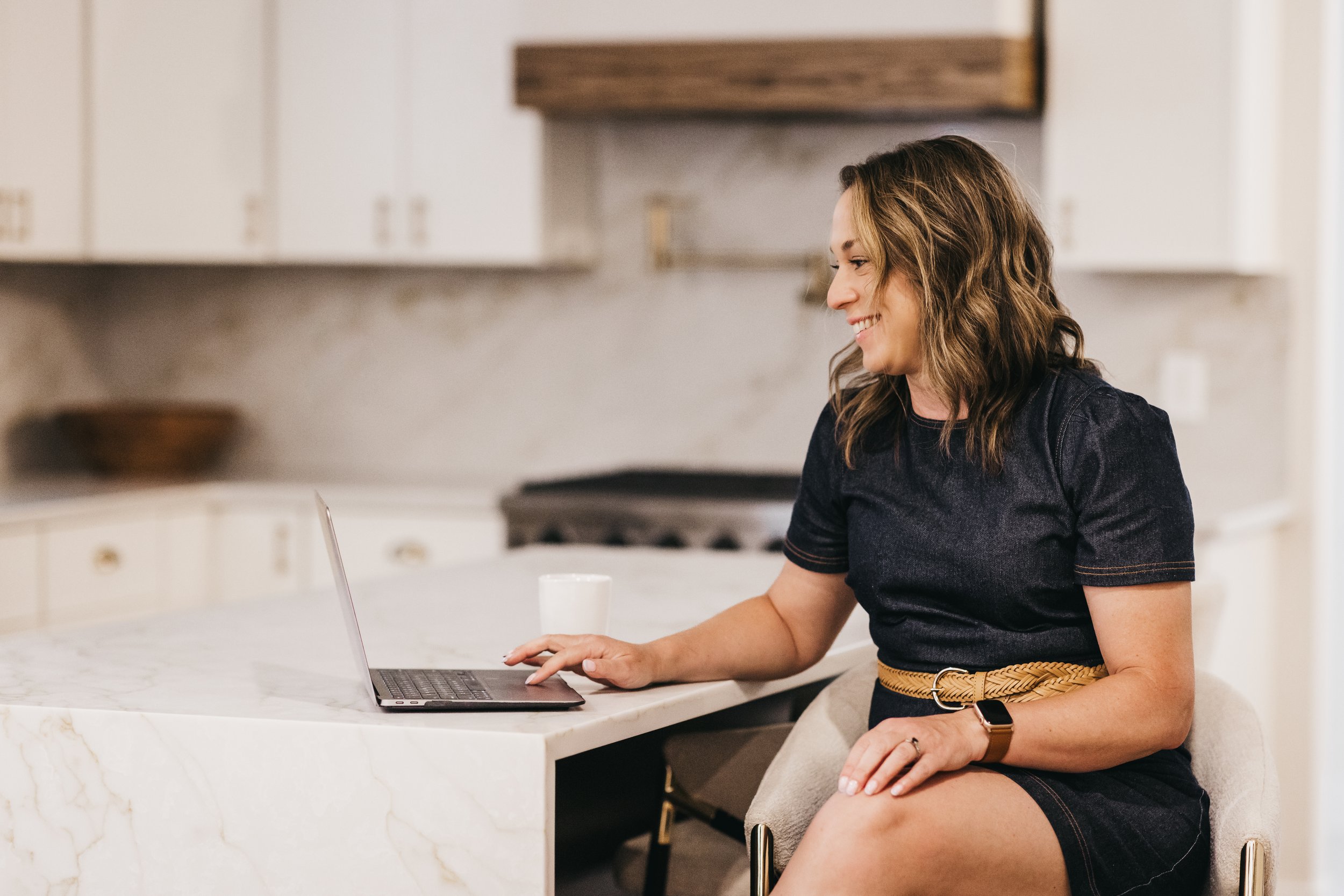 A woman with shoulder-length brown hair sitting in a kitchen, smiling while using a laptop on a white marble kitchen island.