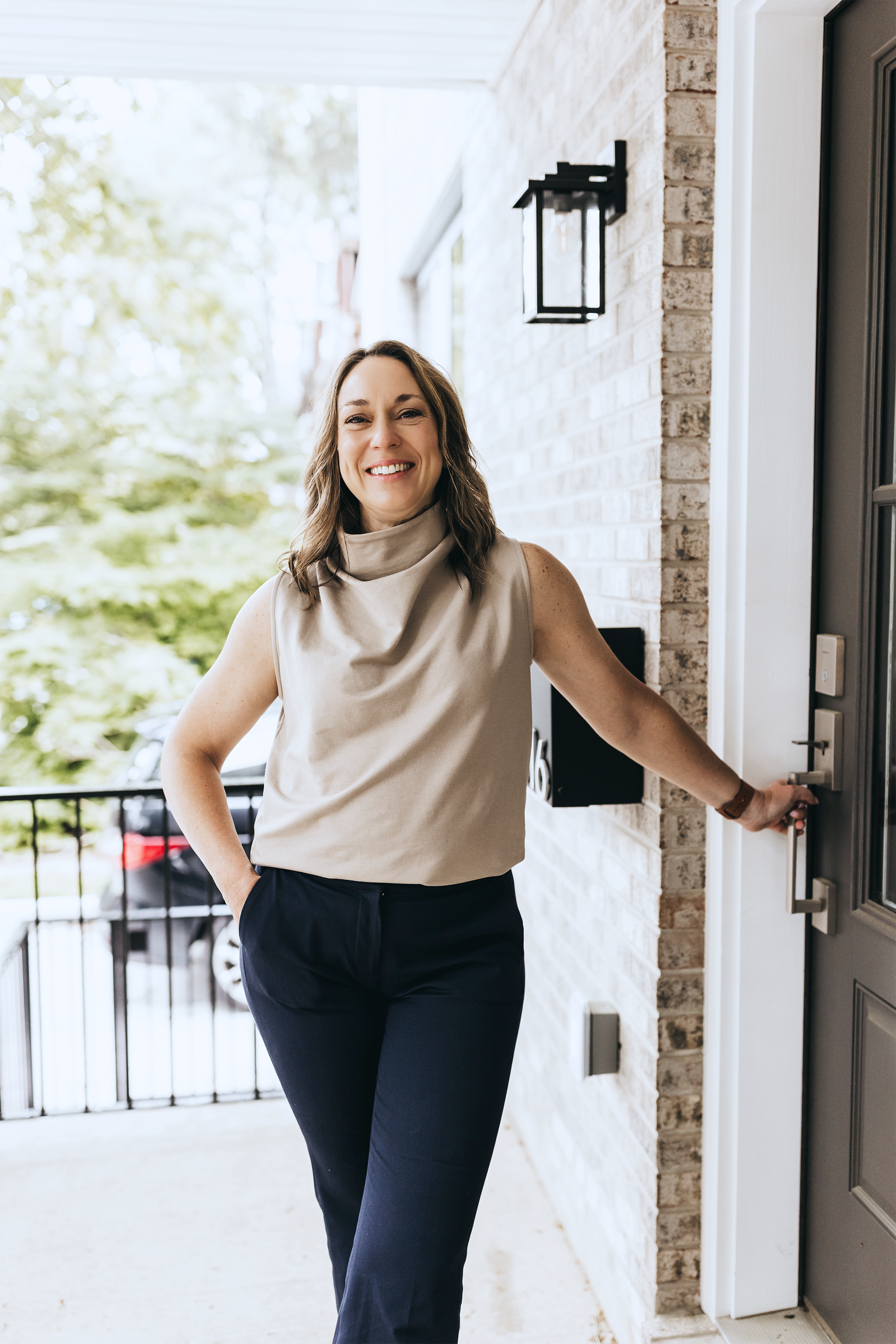 A woman standing at her apartment door, smiling and holding the doorknob, with an outdoor background of trees and parked cars.