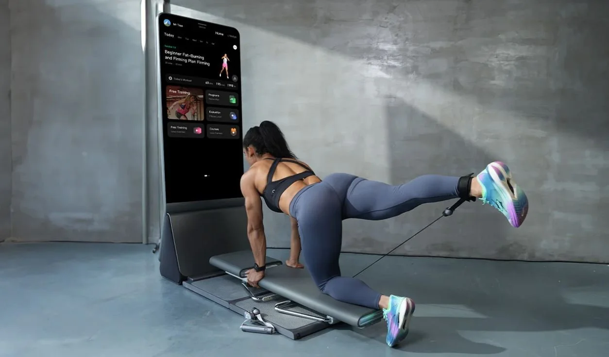 Woman exercising using resistance bands connected to a large touchscreen fitness app in a gym.