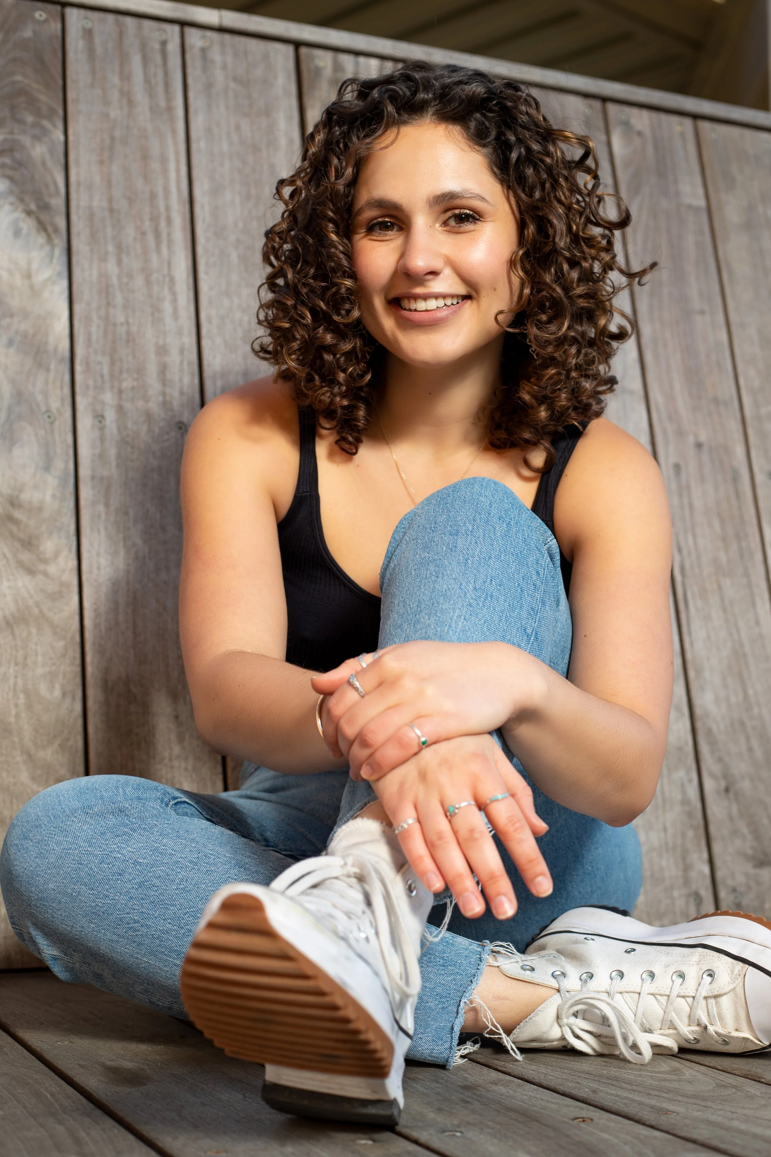 A young woman with curly brown hair sitting on a wooden surface, smiling, wearing a black tank top, blue jeans, and white sneakers, with her hands resting on her knee. BoldVision Studios