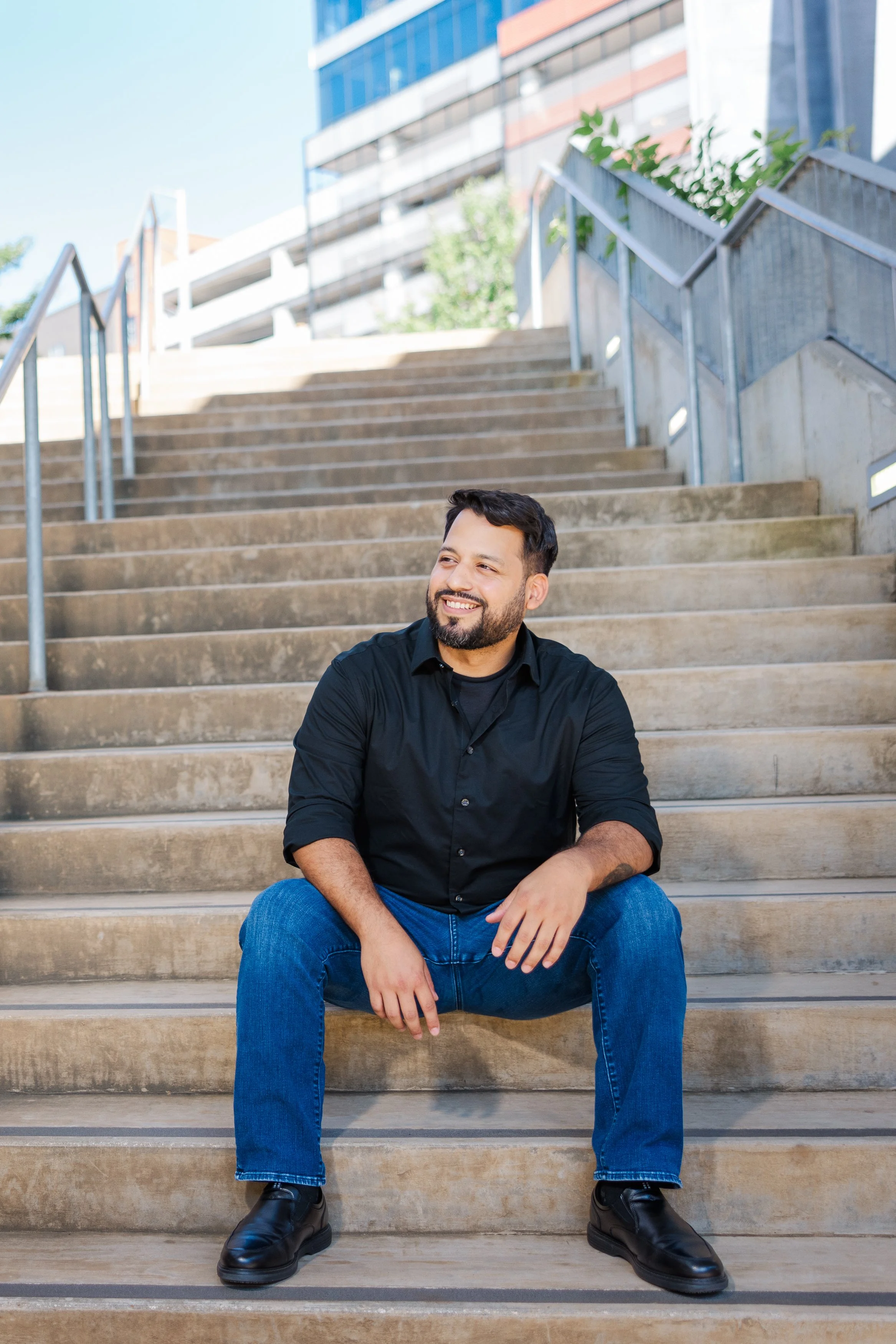 Smiling man in black shirt and jeans sitting on outdoor stairs in an urban setting. BoldVision Studios
