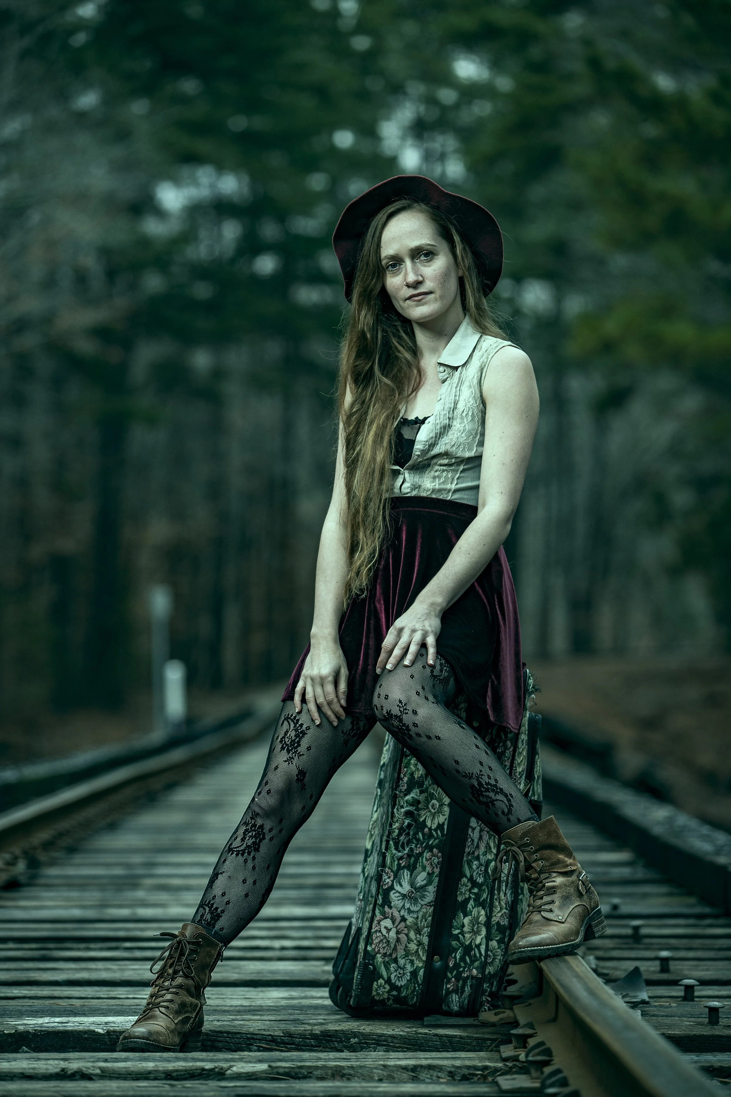 A young woman with long, wavy hair wearing a hat, sleeveless shirt, lace tights, and combat boots, sitting on a suitcase on a railway track in a forest. BoldVision Studios