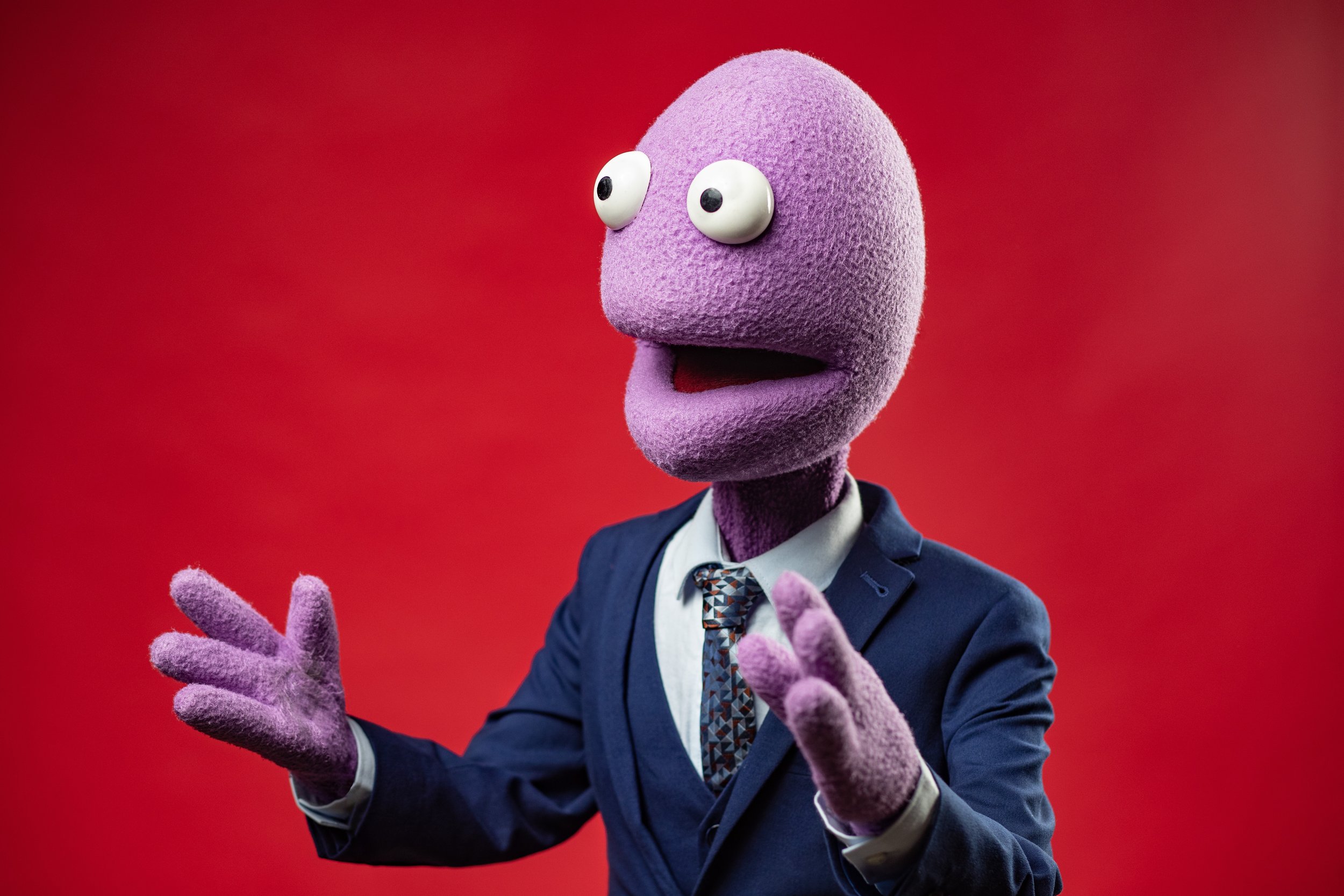 A purple puppet character with large eyes and a wide mouth, dressed in a business suit and tie, gesturing with both hands against a red background. BoldVision Studios